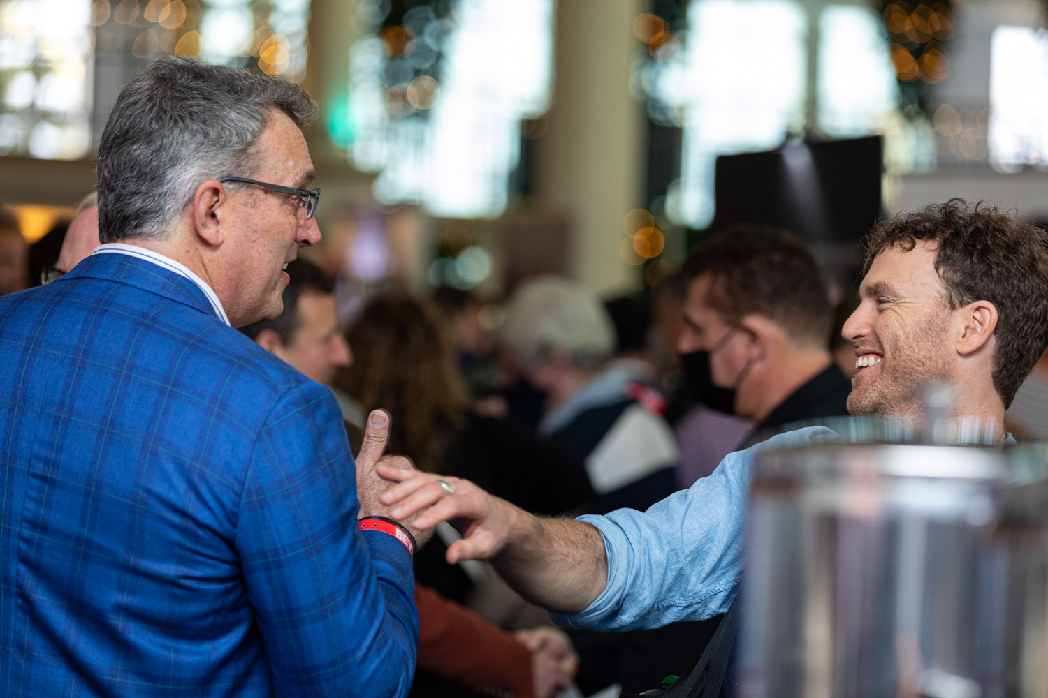 A man in a blue plaid blazer reaches out to shake hands with a curly-haired smiling man, in a crowded room with string lights.