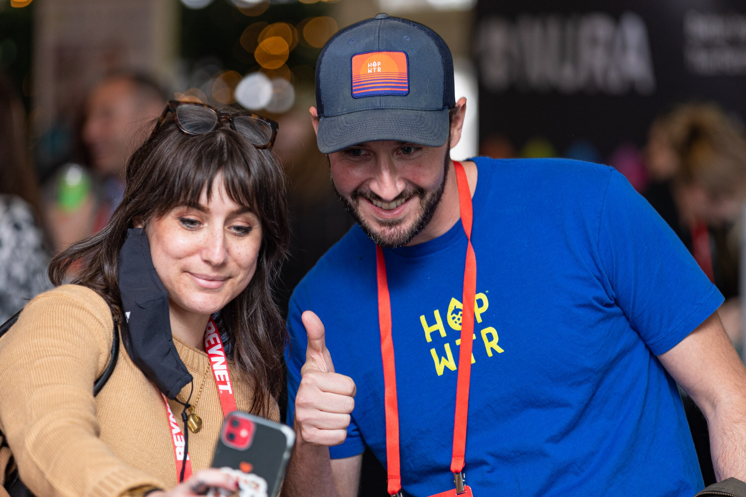A bearded man in a blue Hop Wtr T-shirt and a cap and a woman in a tan top with a mask pulled down smile toward a phone.