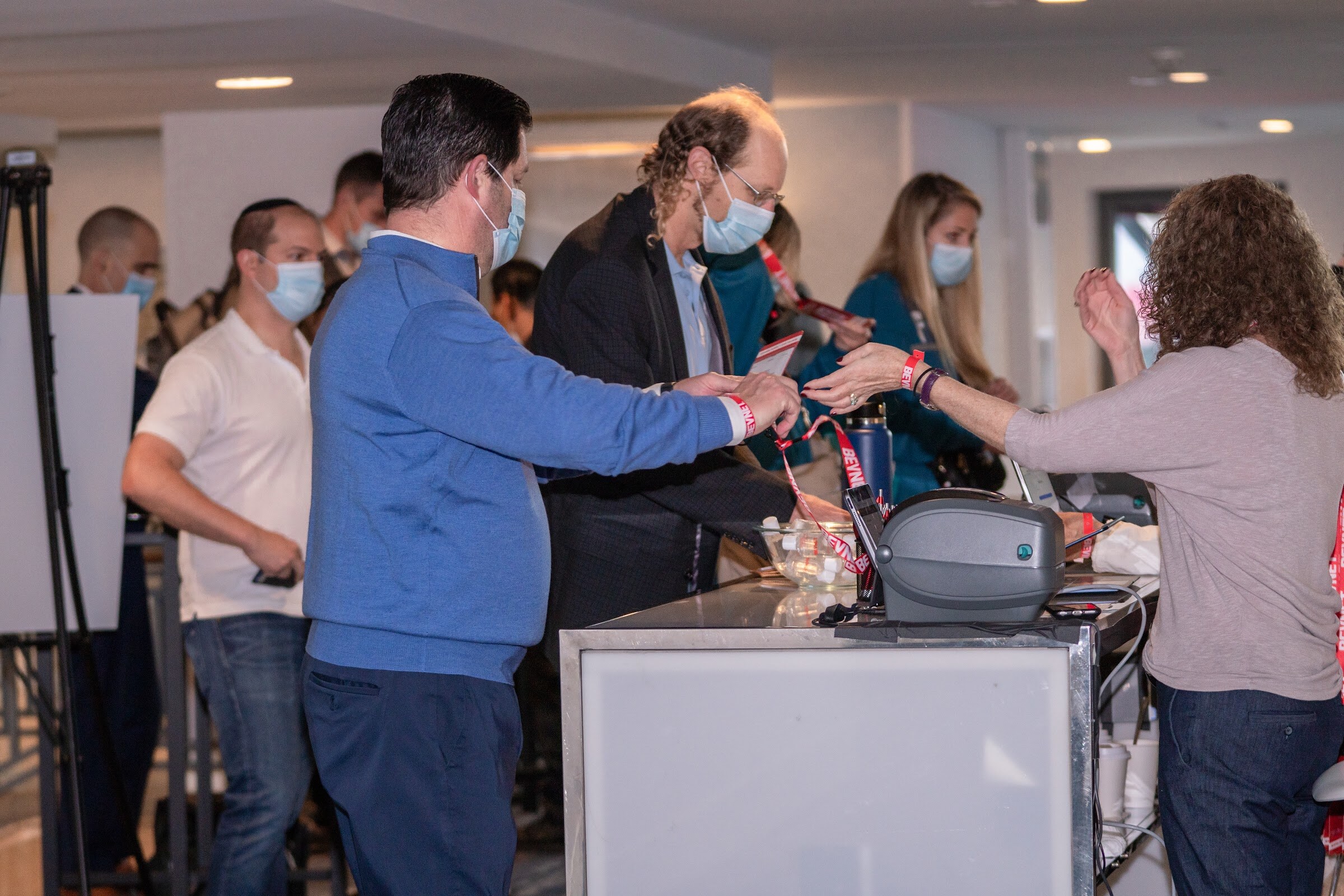 A group of attendees in masks stand at a white registration counter with label printers while a woman in a gray top reaches toward the desk.