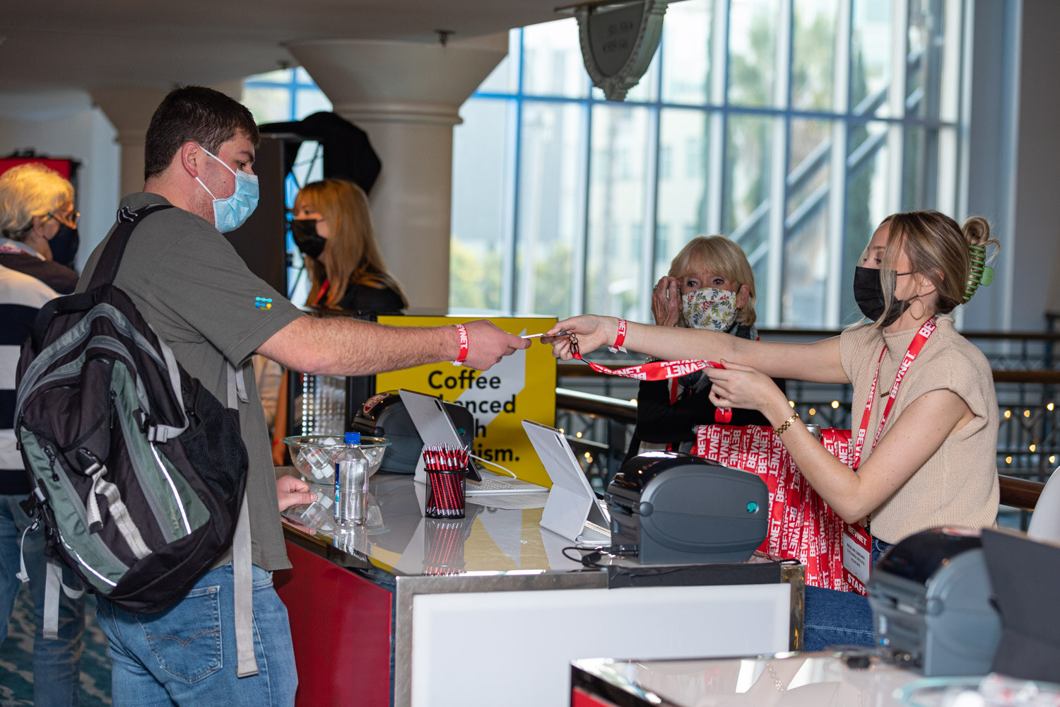 A man with a backpack and mask takes a red lanyard from a woman behind a counter with label printers, a coffee sign and a red bag.