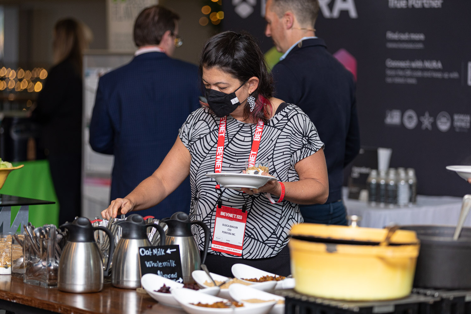 A woman in a patterned top and face mask holds a plate beside coffee pots and serving dishes, with a sign reading Oat Milk on the table.