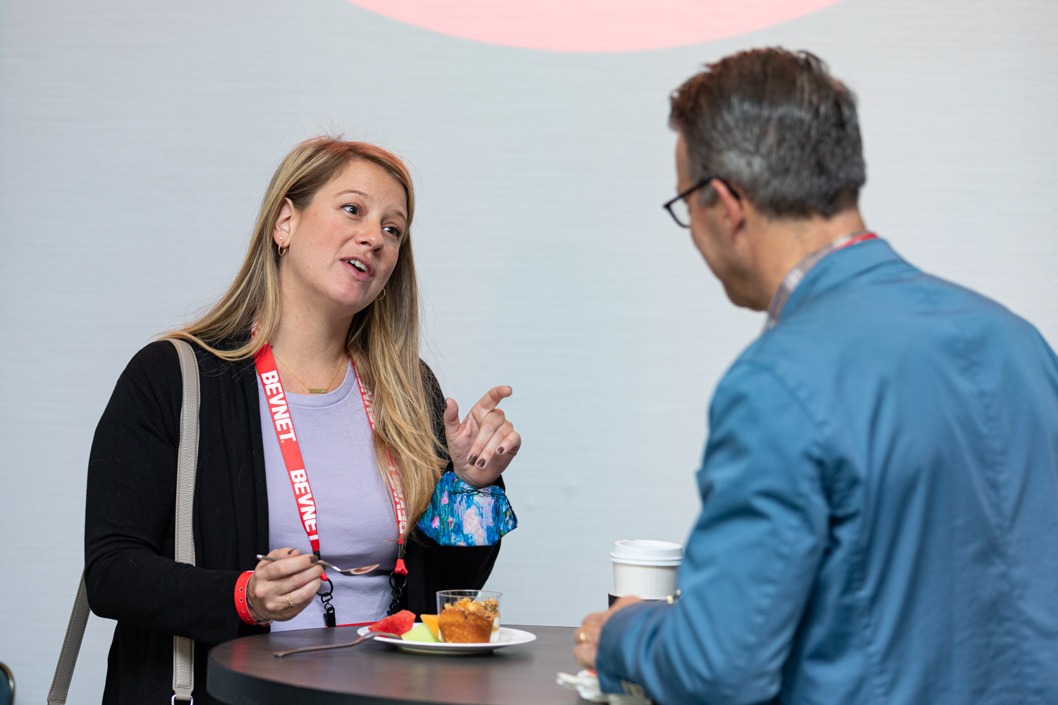 A blonde woman in a black cardigan with a BevNET lanyard talks with a man in a blue jacket across a high-top table with pastries and coffee.