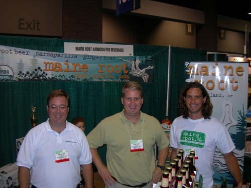Three men stand smiling at a booth with green curtains and Maine Root banners; bottles of soda sit on the counter in front of them.