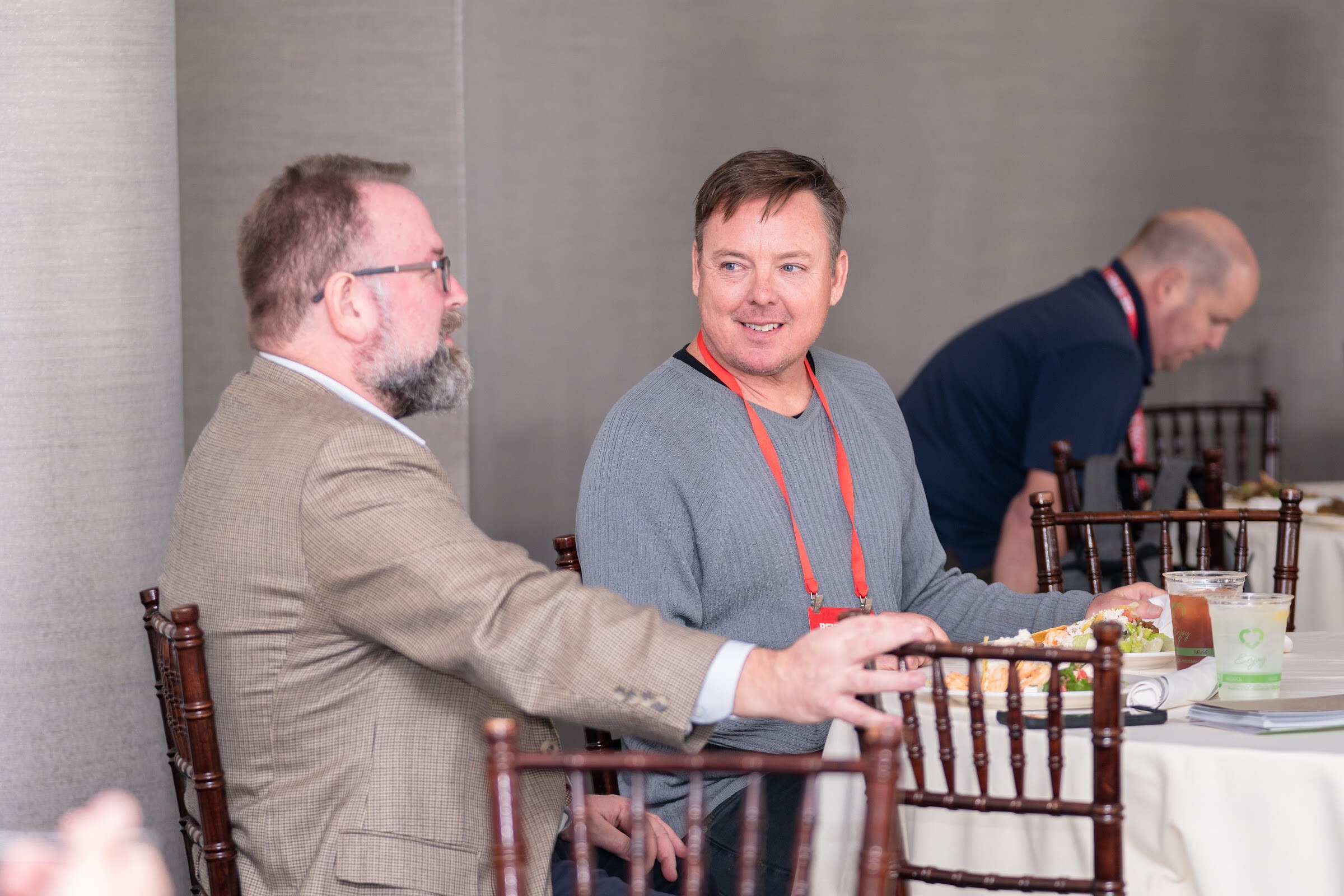 A man in a tan blazer and a smiling man in a gray sweater with a red lanyard sit at a round table with iced drinks.