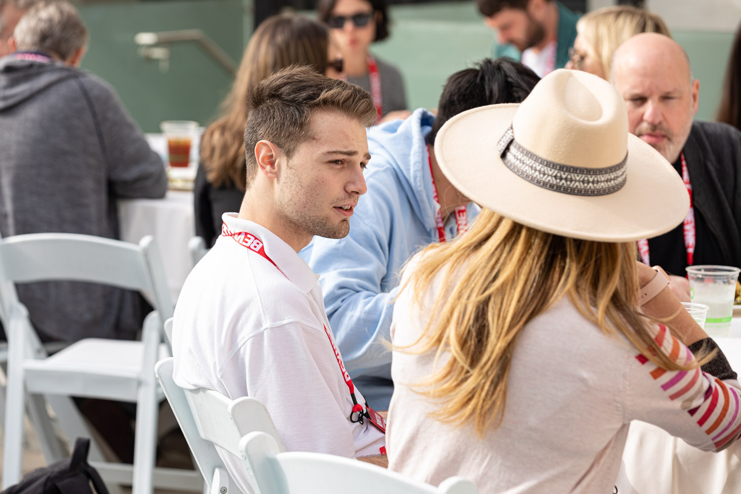 A group seated at an outdoor table, with a woman in a cream wide-brimmed hat in the foreground facing a young man in a white shirt and BevNET lanyard.