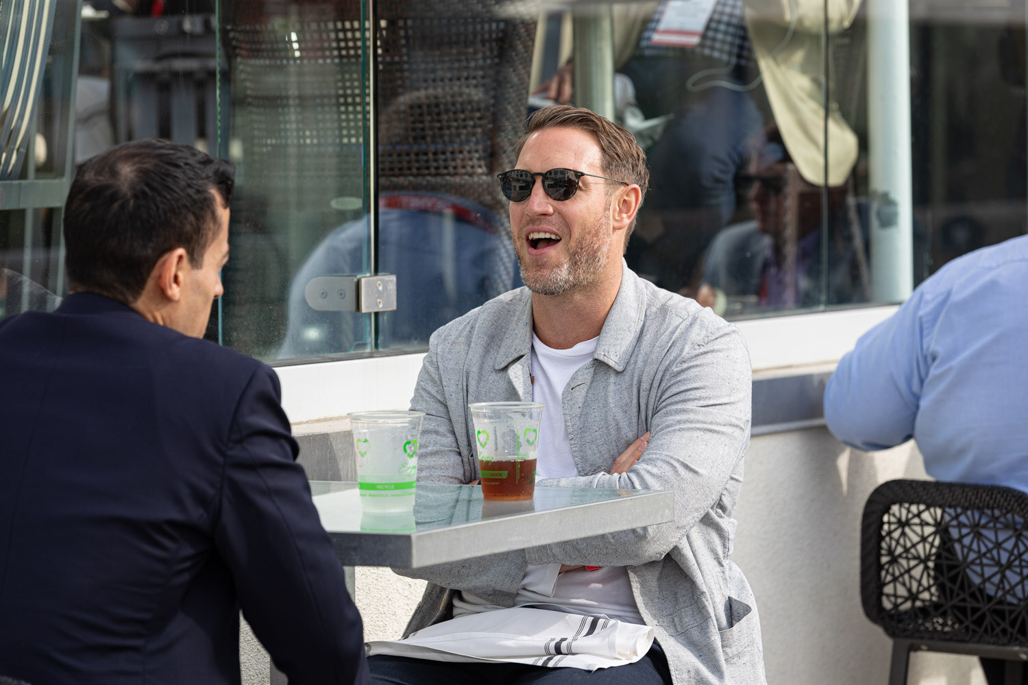 A man in sunglasses and a gray jacket smiles across a small table with iced teas from a man in a dark blazer seen from behind.