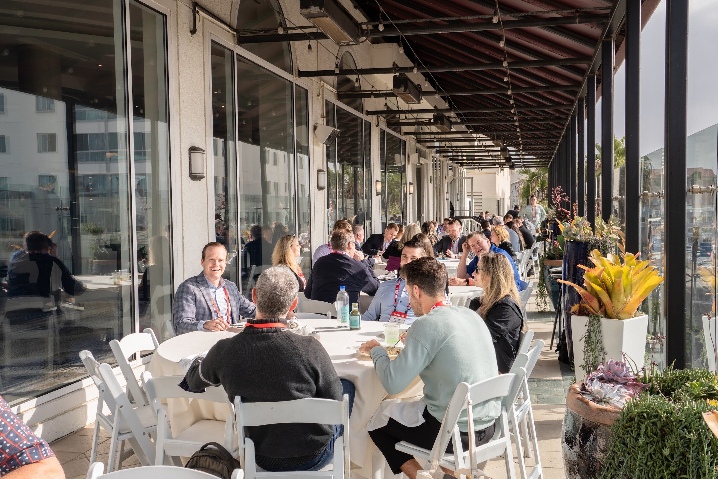A long covered terrace with attendees seated at round tables with white folding chairs, large windows on the left and potted plants at right.