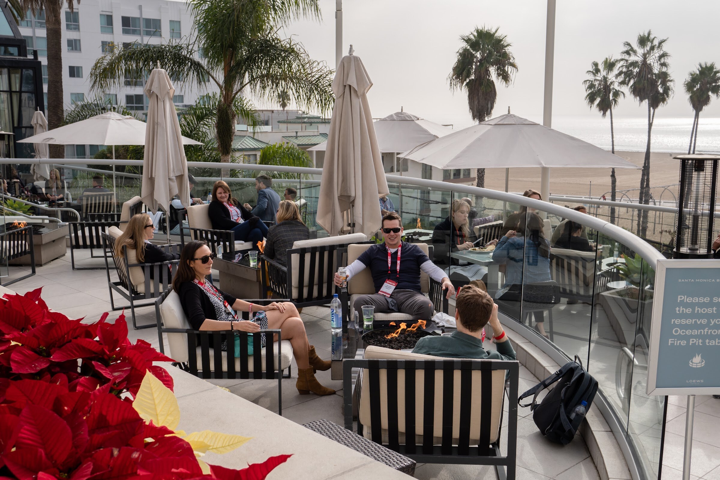 Groups sit in cushioned chairs around fire pit tables on a curved terrace with palm trees and beach umbrellas beyond a glass railing.