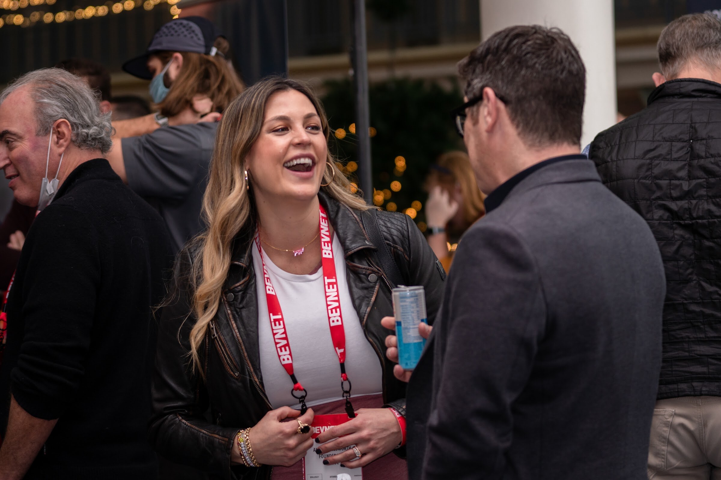 A woman with long wavy hair in a leather jacket and red BevNET lanyard holds a can and laughs while talking with others.