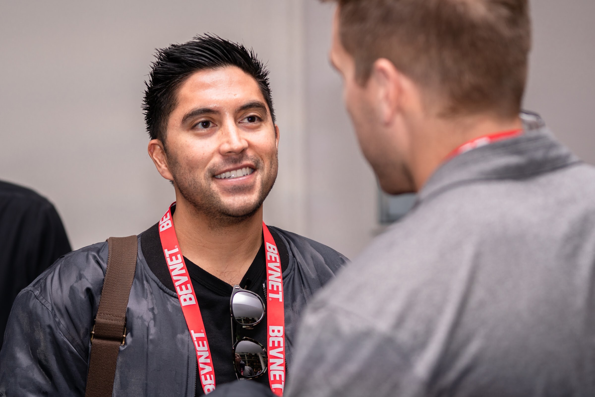 A dark-haired man in a bomber jacket with a BevNET lanyard smiles while facing a man seen from behind.