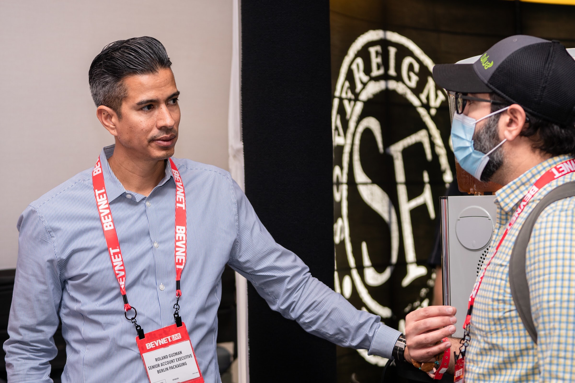 A man in a light blue shirt with a BevNET badge reaches out his hand toward a masked man in a cap and checked shirt, in front of a Sovereign Flavors logo.