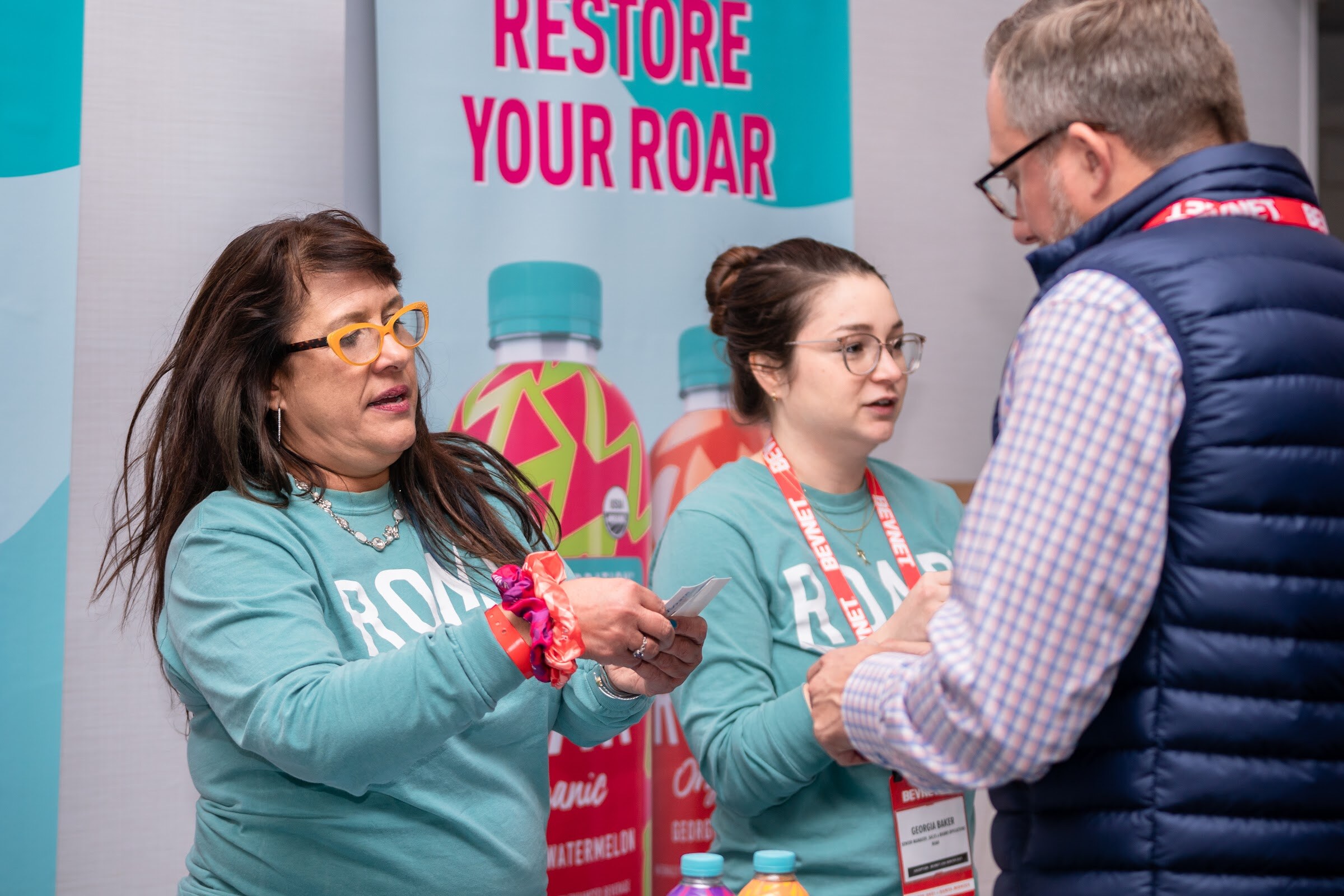 A woman in glasses and a teal Roar sweatshirt hands something to a man in a navy puffer vest, with a second teal-sweatshirt woman beside her.