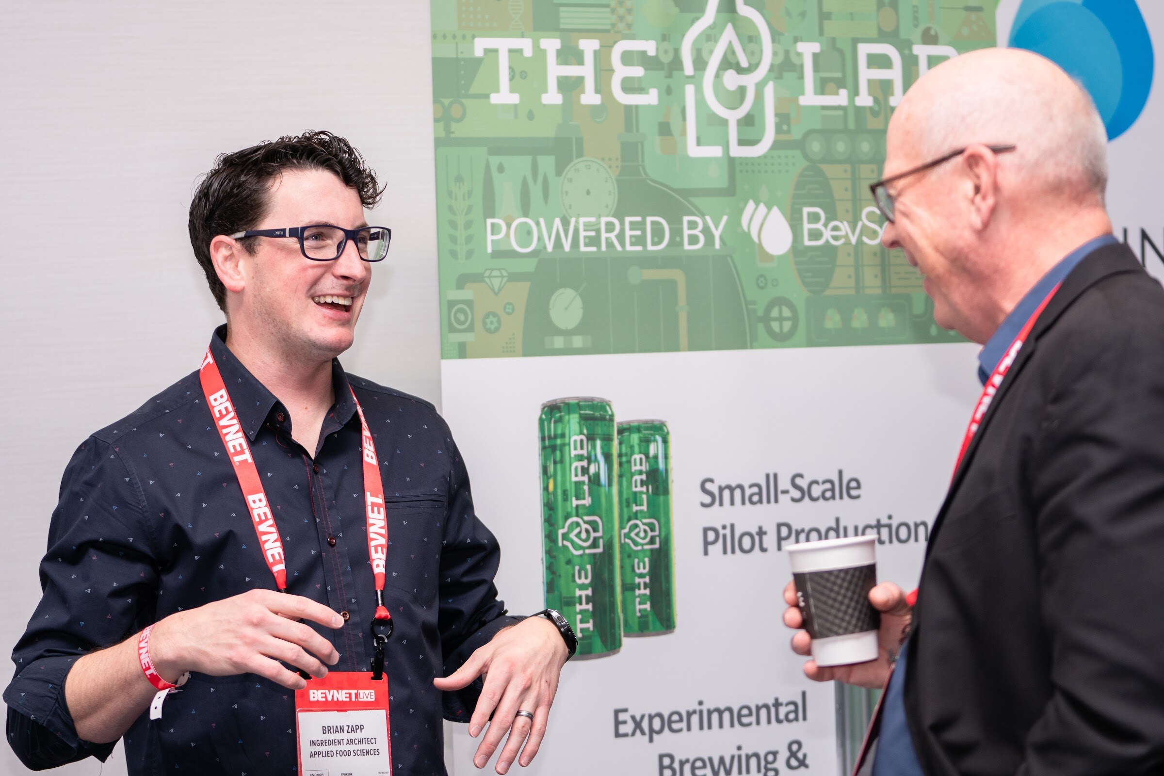A man with glasses and a navy shirt with a BevNET lanyard smiles at a man holding a coffee cup in front of a The Lab Powered by BevSource banner.
