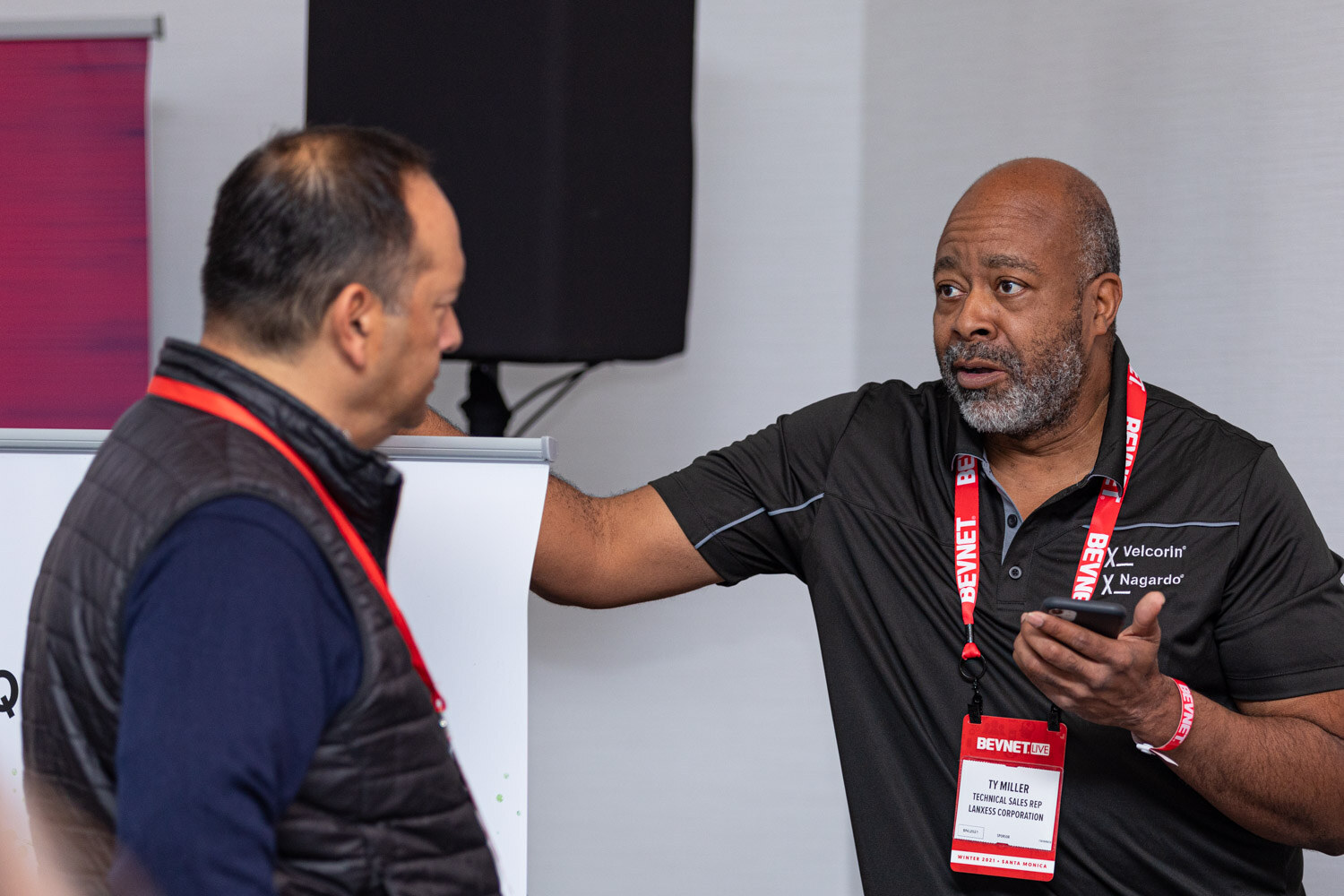 A man with a gray beard in a black polo and a red BevNET lanyard holds a phone and gestures toward a man in a dark vest in a room with a flip chart.