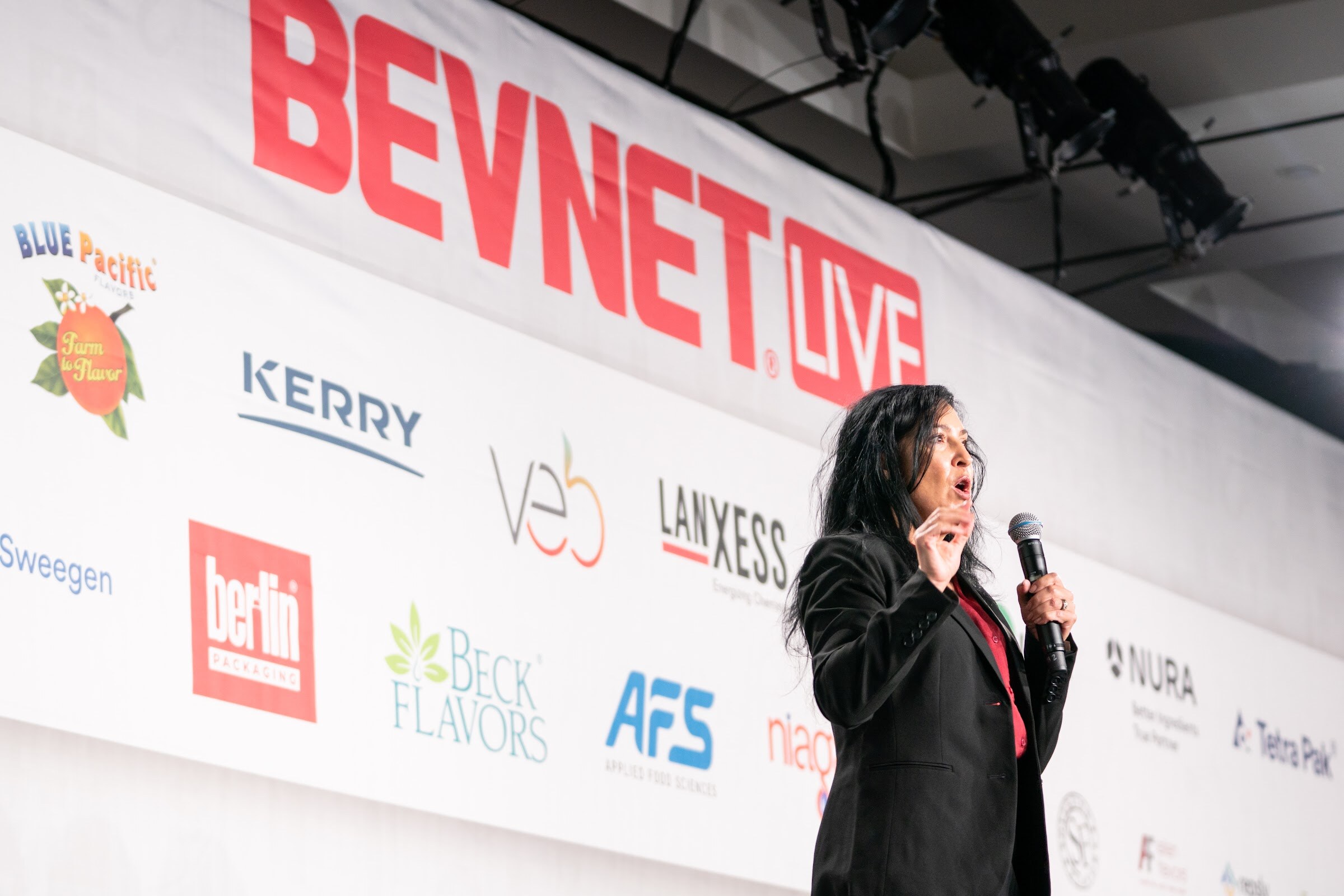A woman with long dark hair in a black blazer speaks into a handheld microphone beside a BevNET Live banner and sponsor logos.