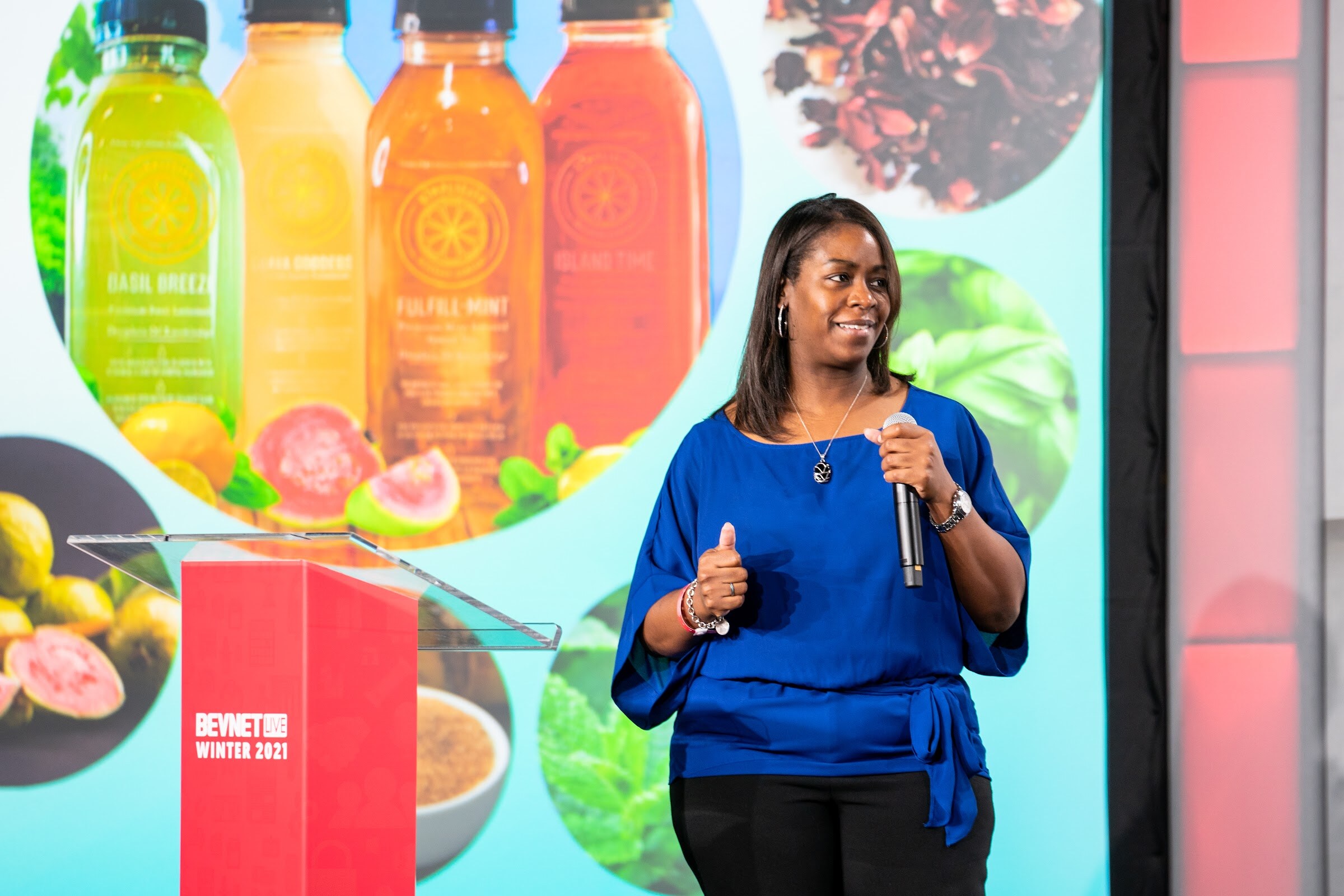 A woman in a bright blue top holds a microphone beside a red BevNET podium with a slide of bottled drinks and fruit behind her.