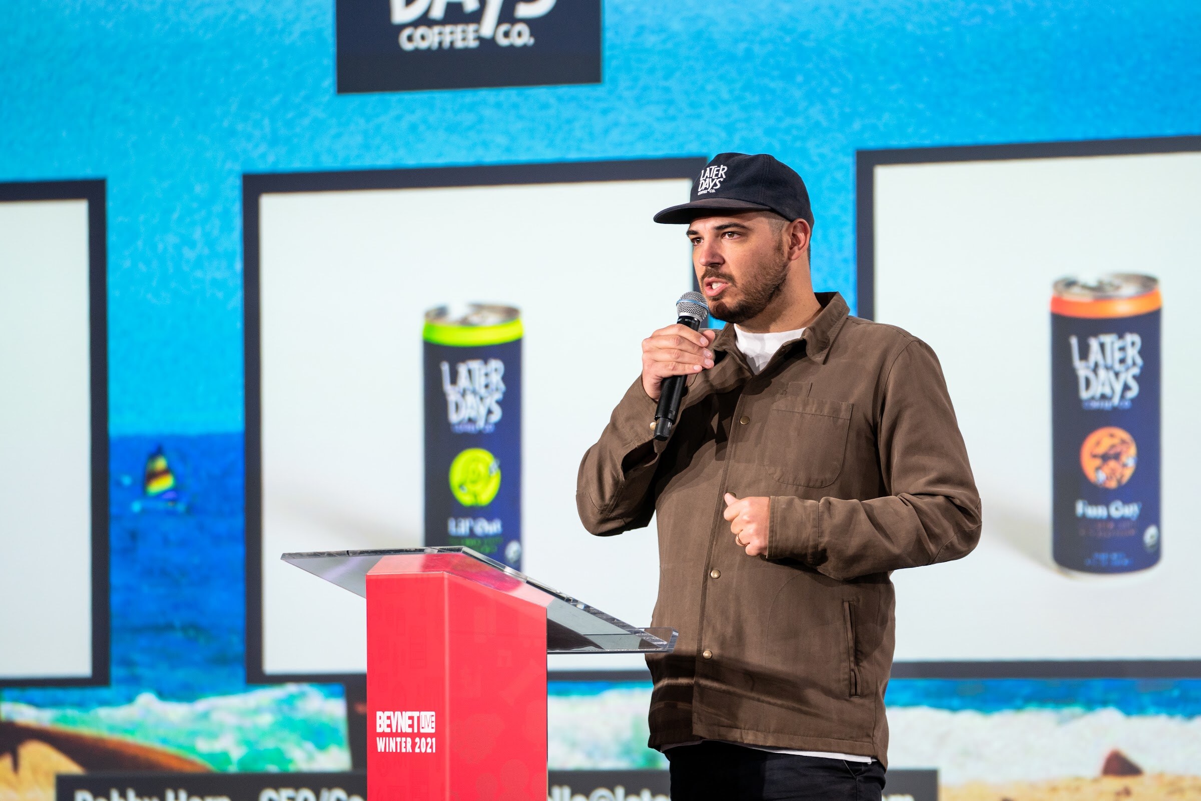 A man in a brown jacket and a Later Days cap speaks into a microphone in front of a slide showing two Later Days cans.