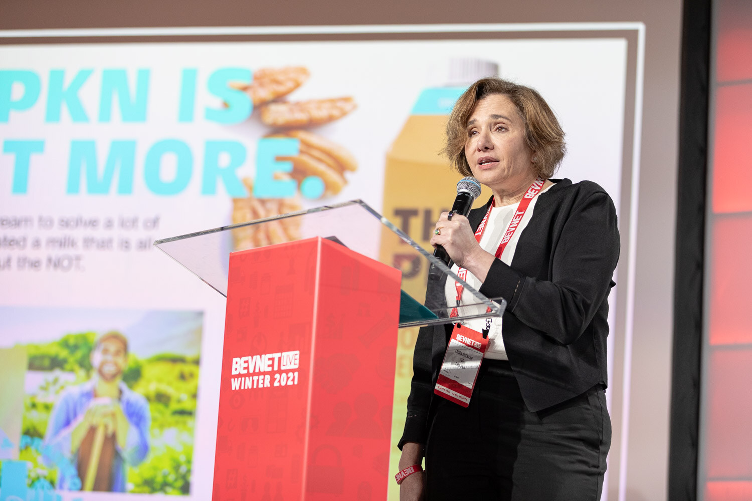 A woman with a BevNET lanyard speaks into a microphone at a red podium in front of a slide reading PKN is more and a photo of pecans.