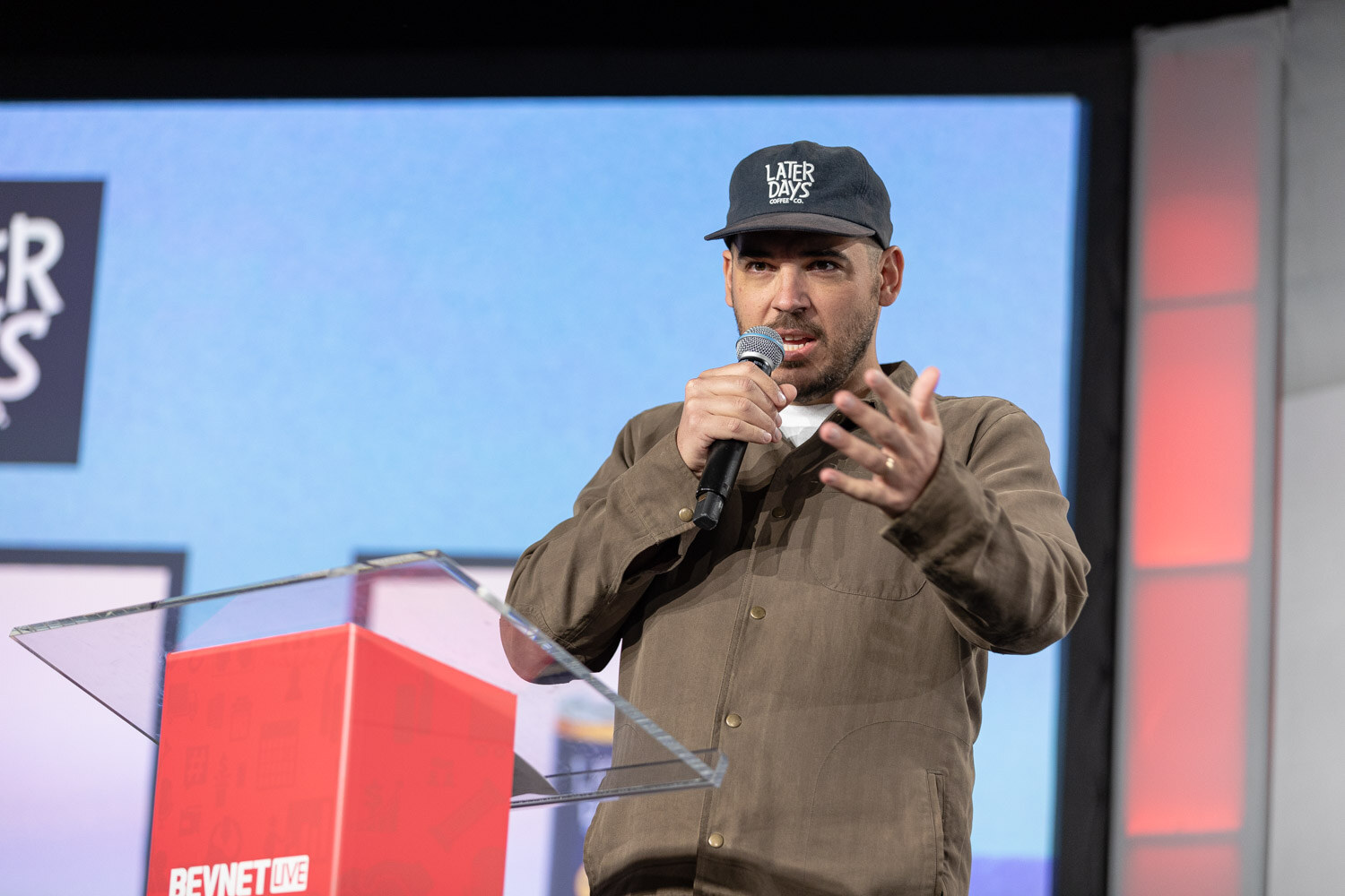 A man in a brown jacket and a navy Later Days cap speaks into a microphone with one hand raised, in front of a bright blue slide and a red podium.
