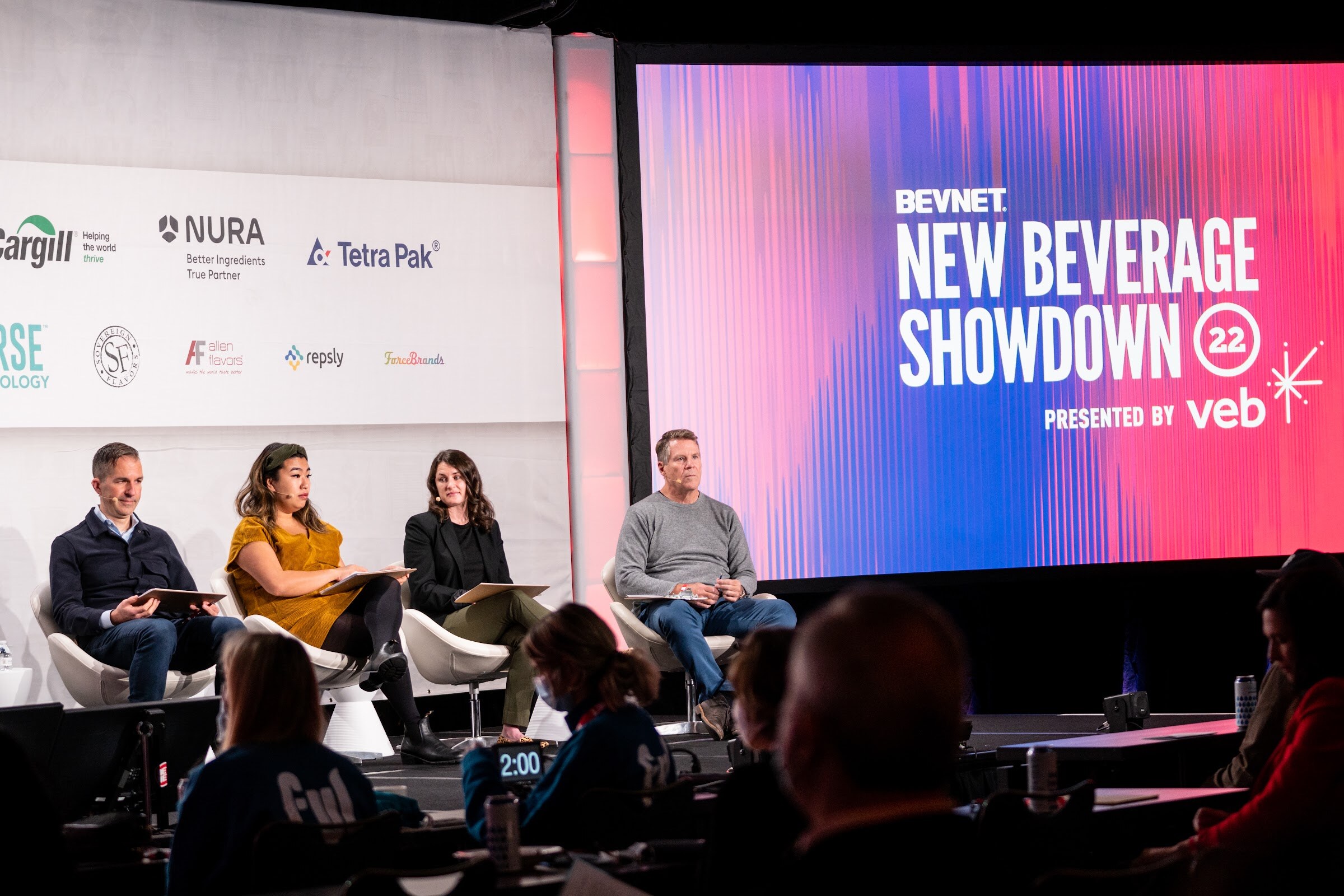 Four judges seated with clipboards on a stage next to a large screen reading BevNET New Beverage Showdown 22 presented by veb.