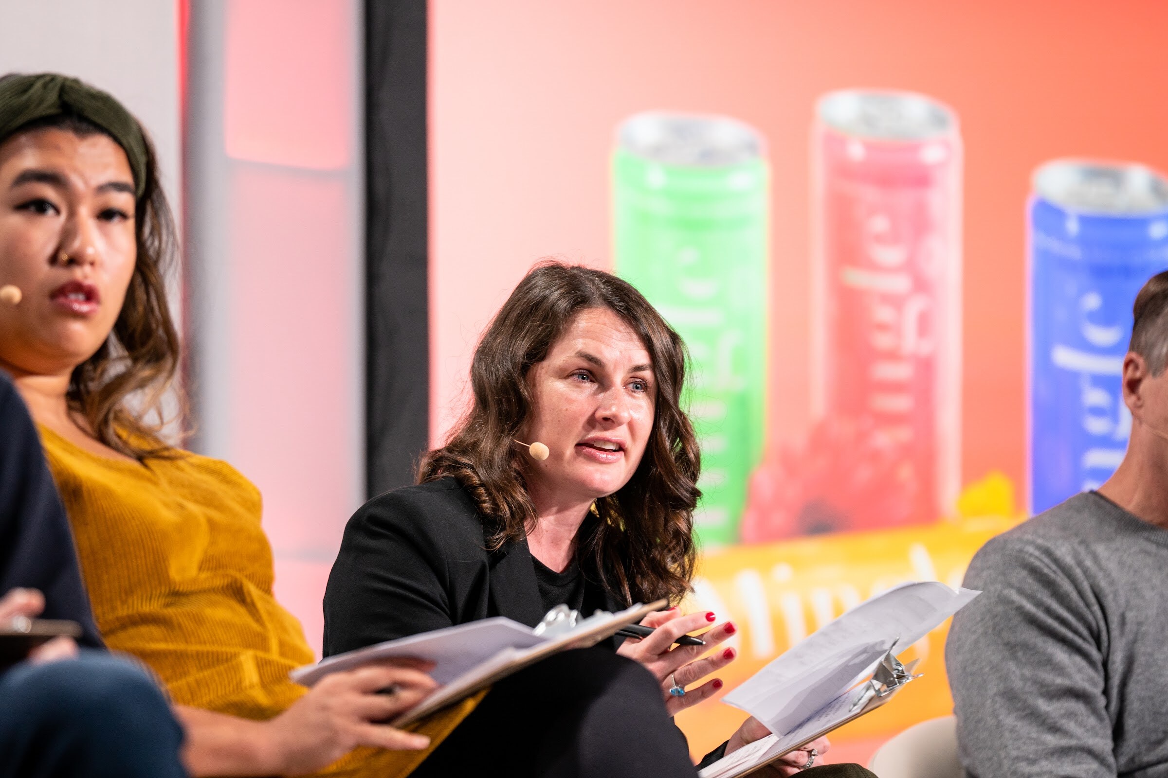 A woman in a mustard top and a woman in a black blazer holding papers sit in front of a slide showing colorful cans.