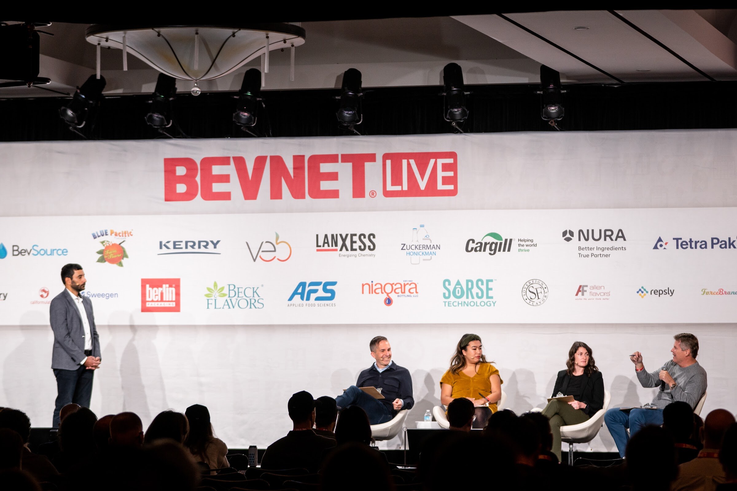 A wide view of a stage under a BevNET Live banner with a man standing at left and four judges seated in front of a seated audience.