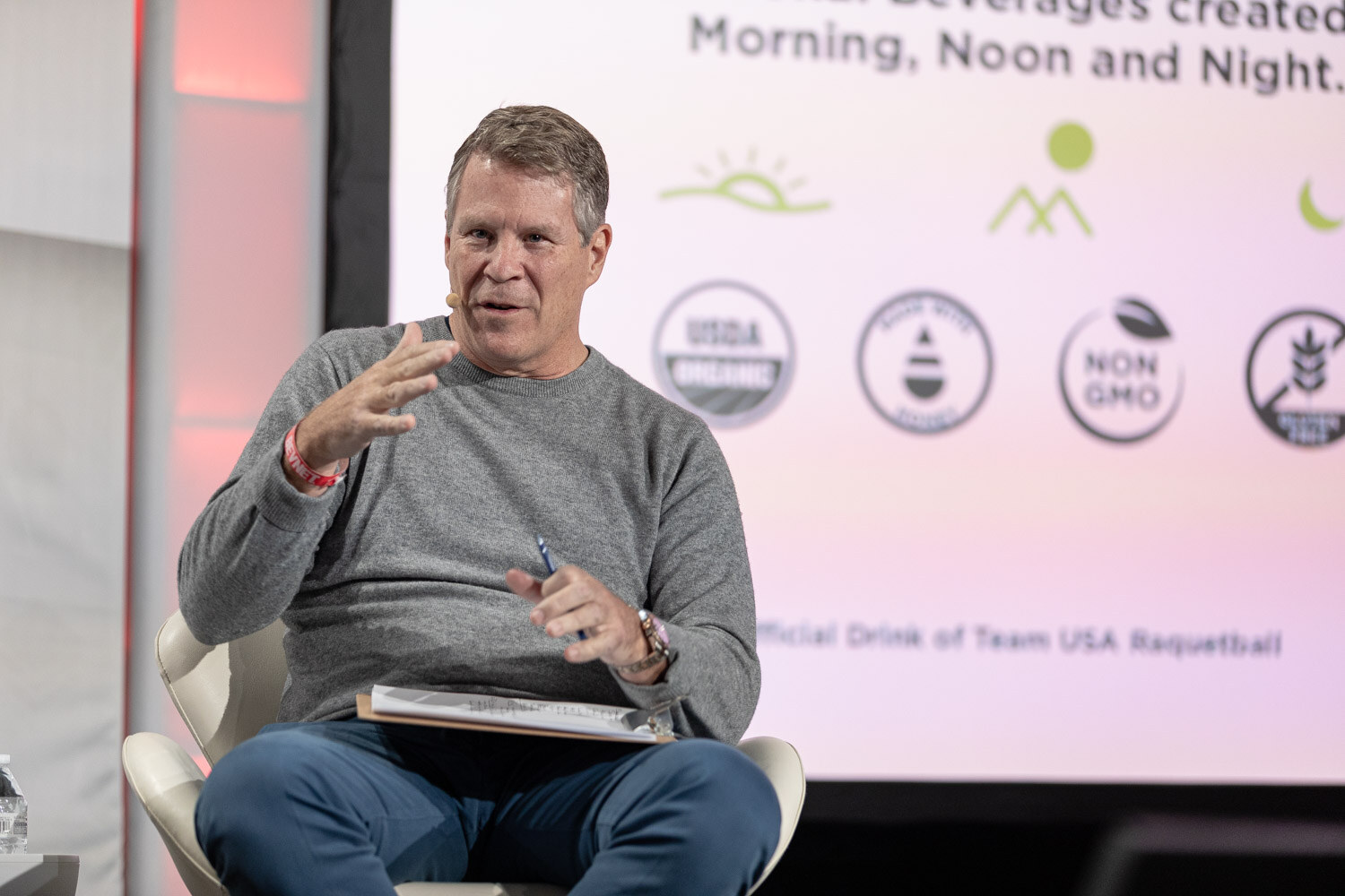 A man in a gray sweater with a clipboard on his lap gestures with one hand in front of a slide with product claims such as USDA Organic and Non GMO.