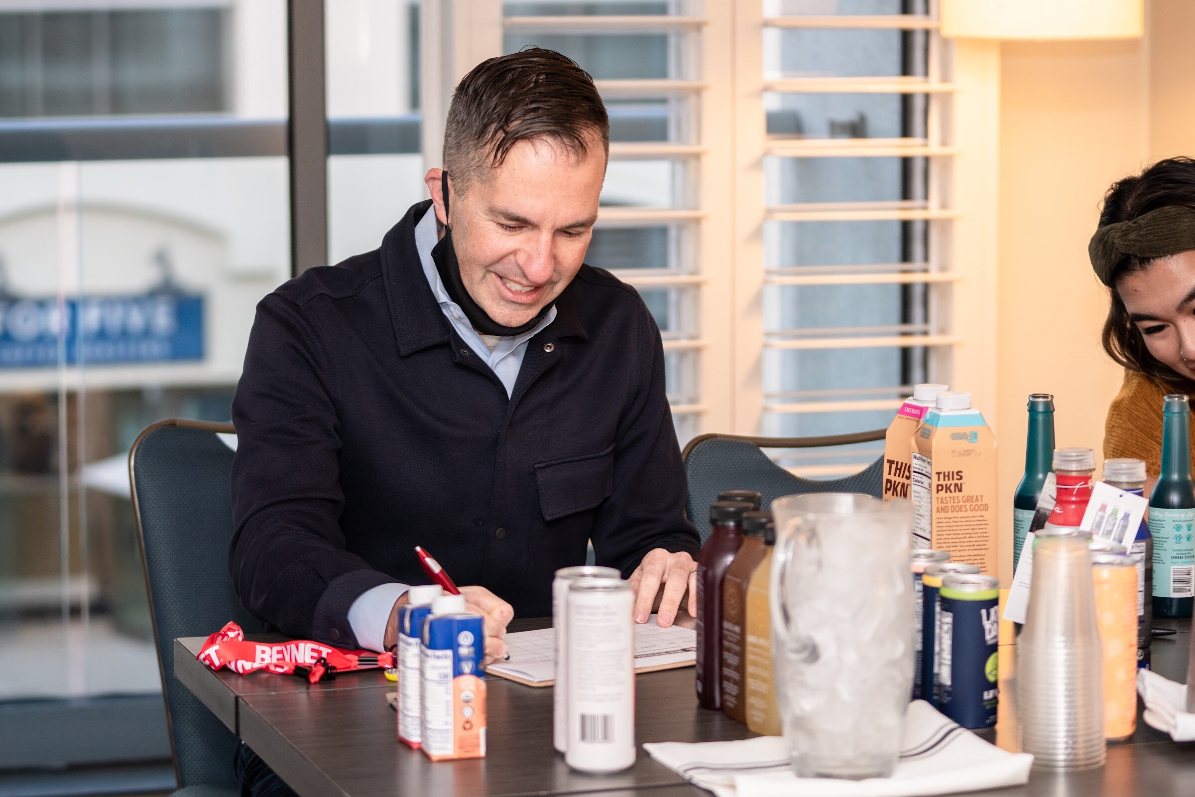 A man in a dark jacket looks down at a score sheet at a table with cartons, cans, bottles and a pitcher of ice.