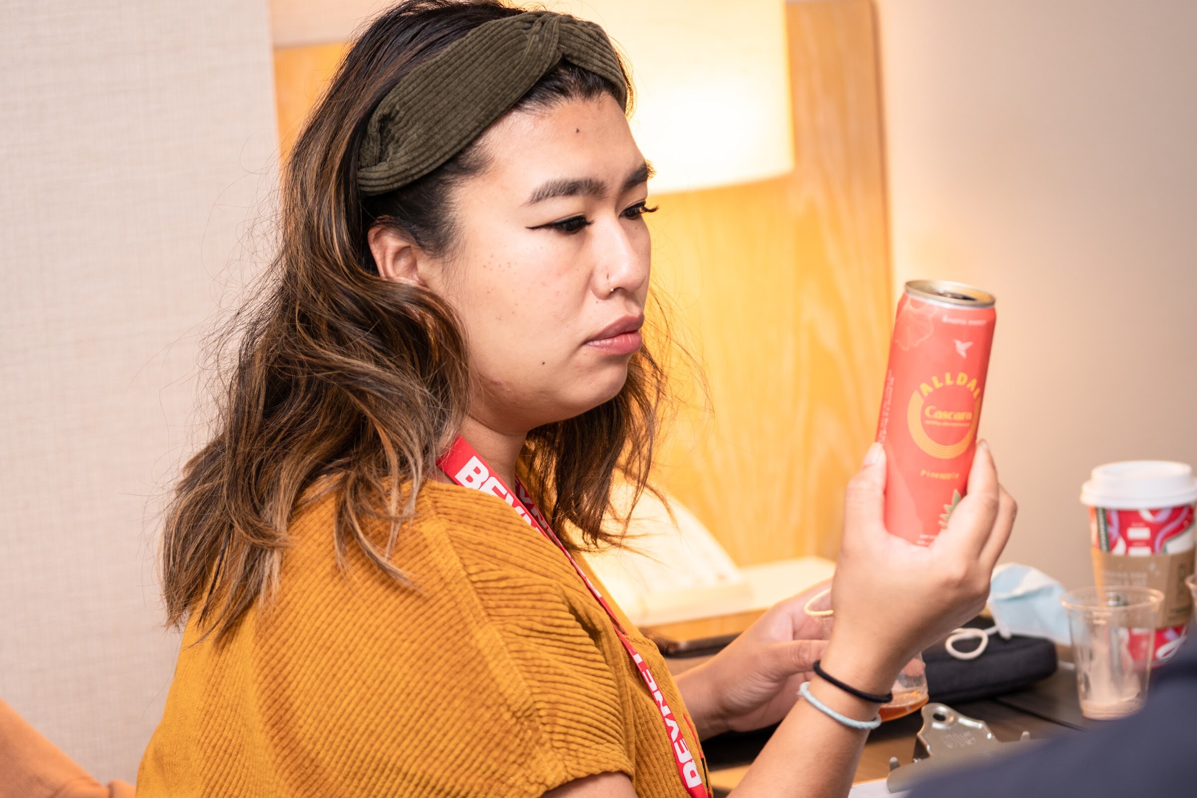 A woman in a mustard top and BevNET lanyard looks at a coral can she is holding up in a hotel room.