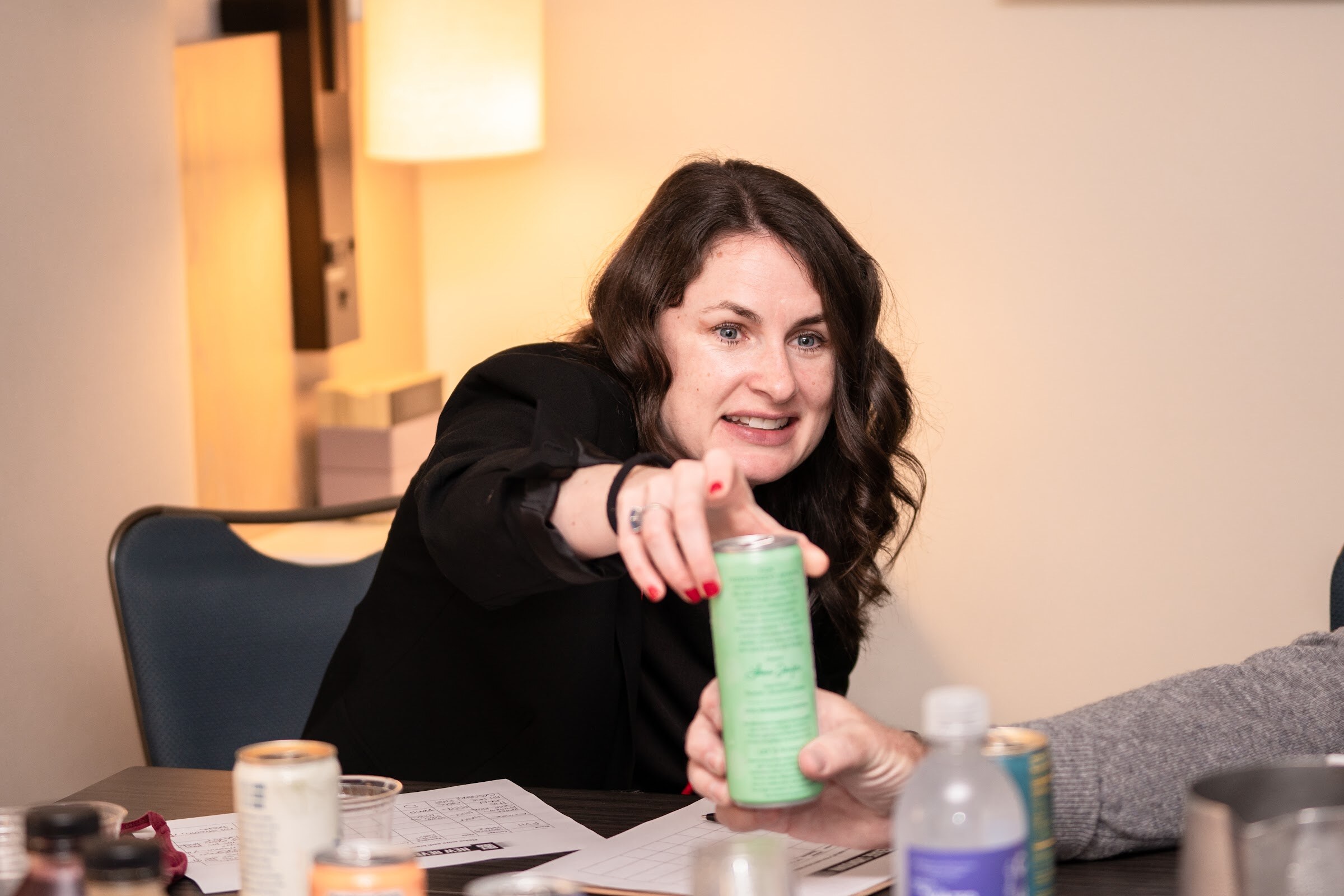 A woman with dark wavy hair in a black top reaches across a table with a green can in her hand, with other cans and cups in front of her.