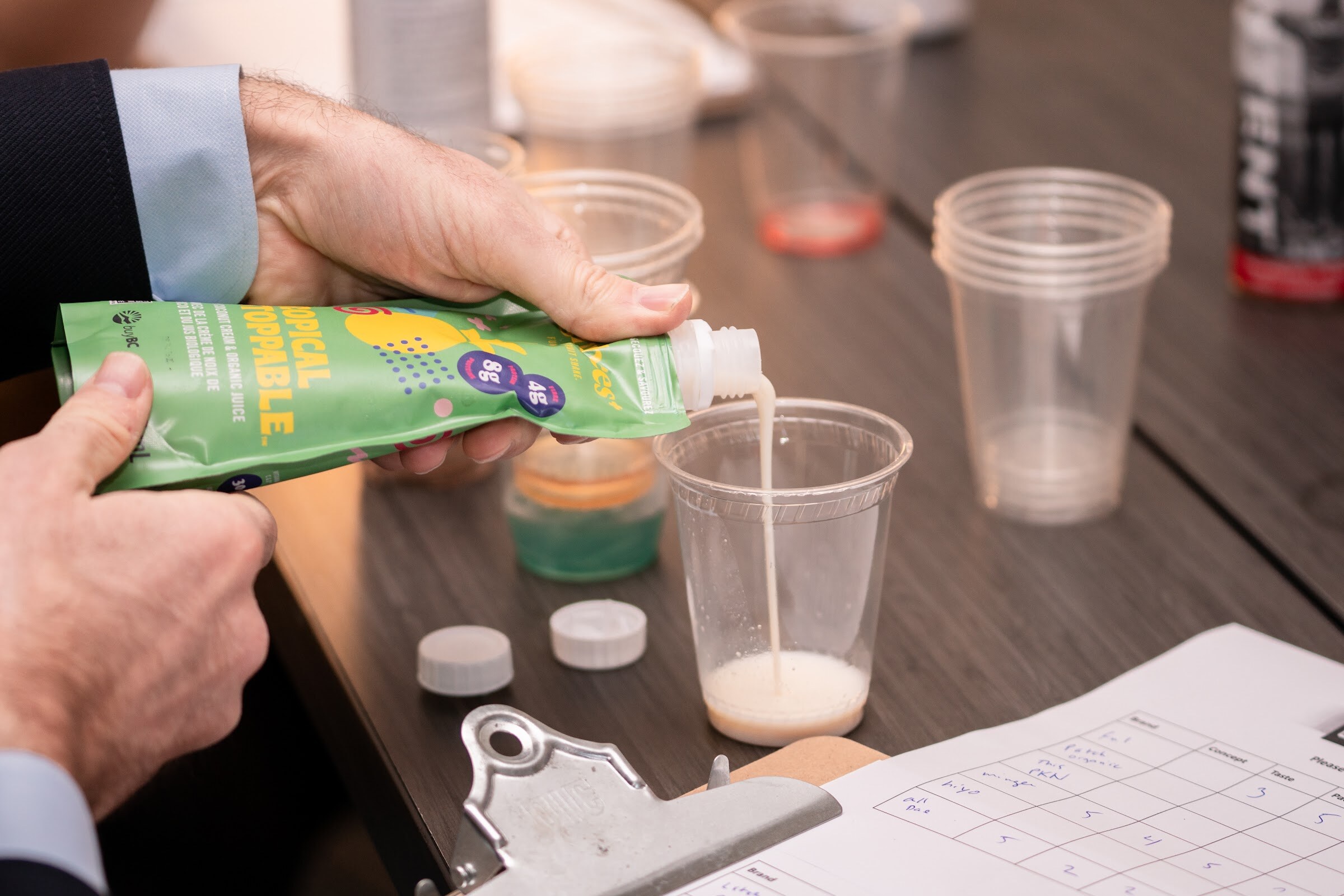 A hand tips a green pouch labeled Tropical into a clear plastic cup next to a score sheet on a clipboard.
