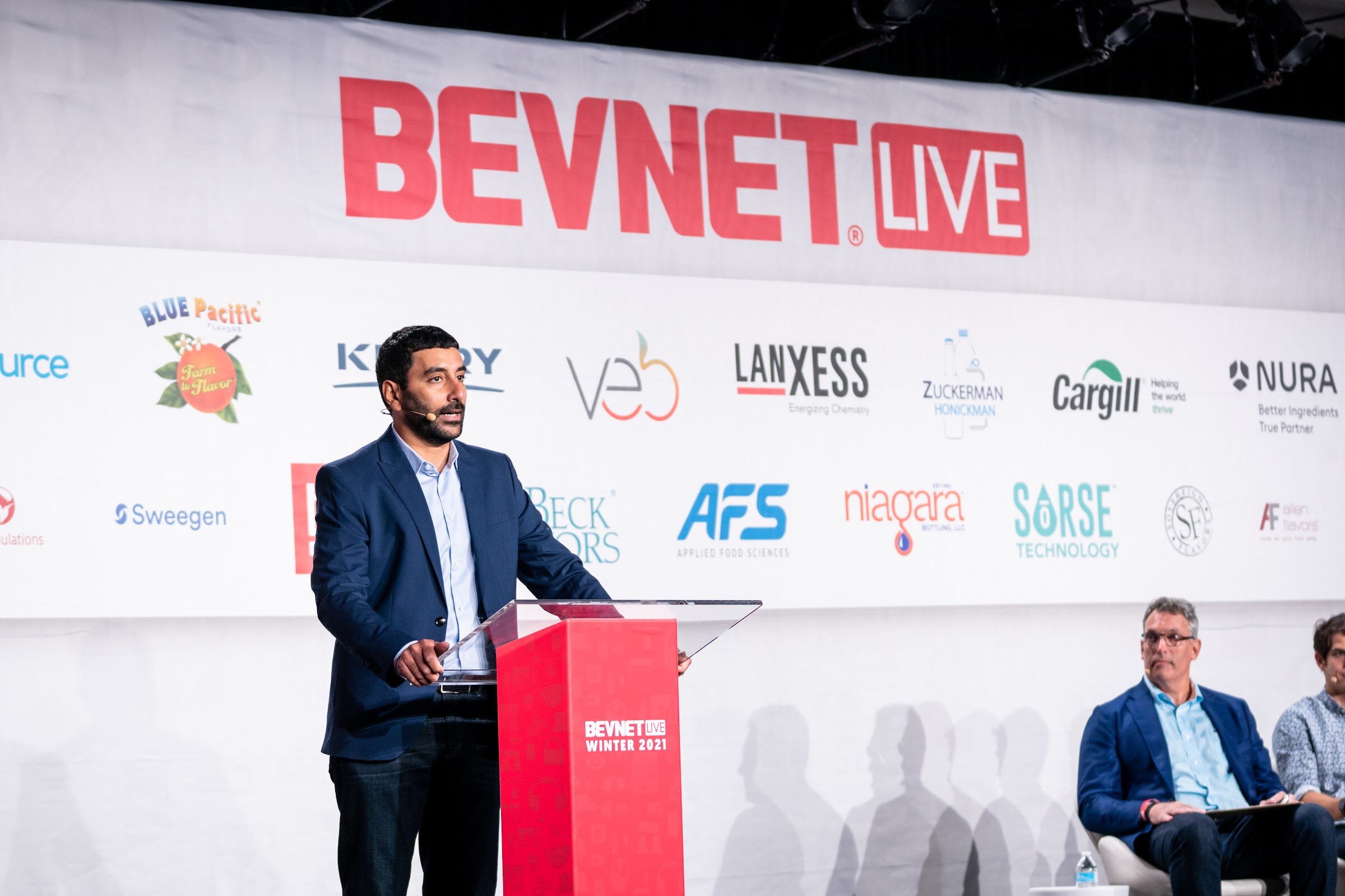 A man in a navy blazer stands at a red podium before a BevNET Live banner and sponsor logos, with two judges seated at the right edge of the stage.