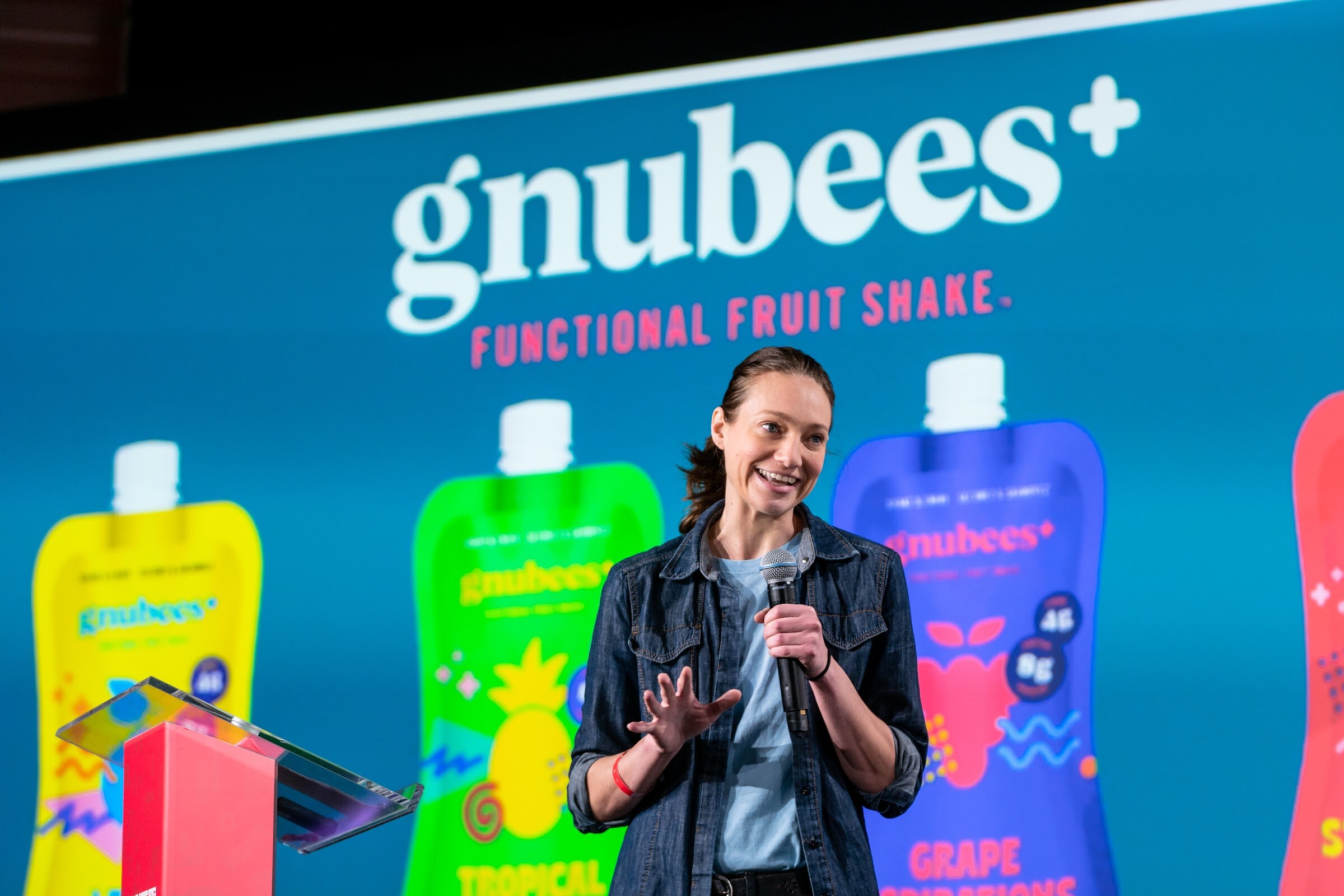 A smiling woman in a denim jacket speaks into a microphone in front of a large slide reading gnubees functional fruit shake with colorful pouches.