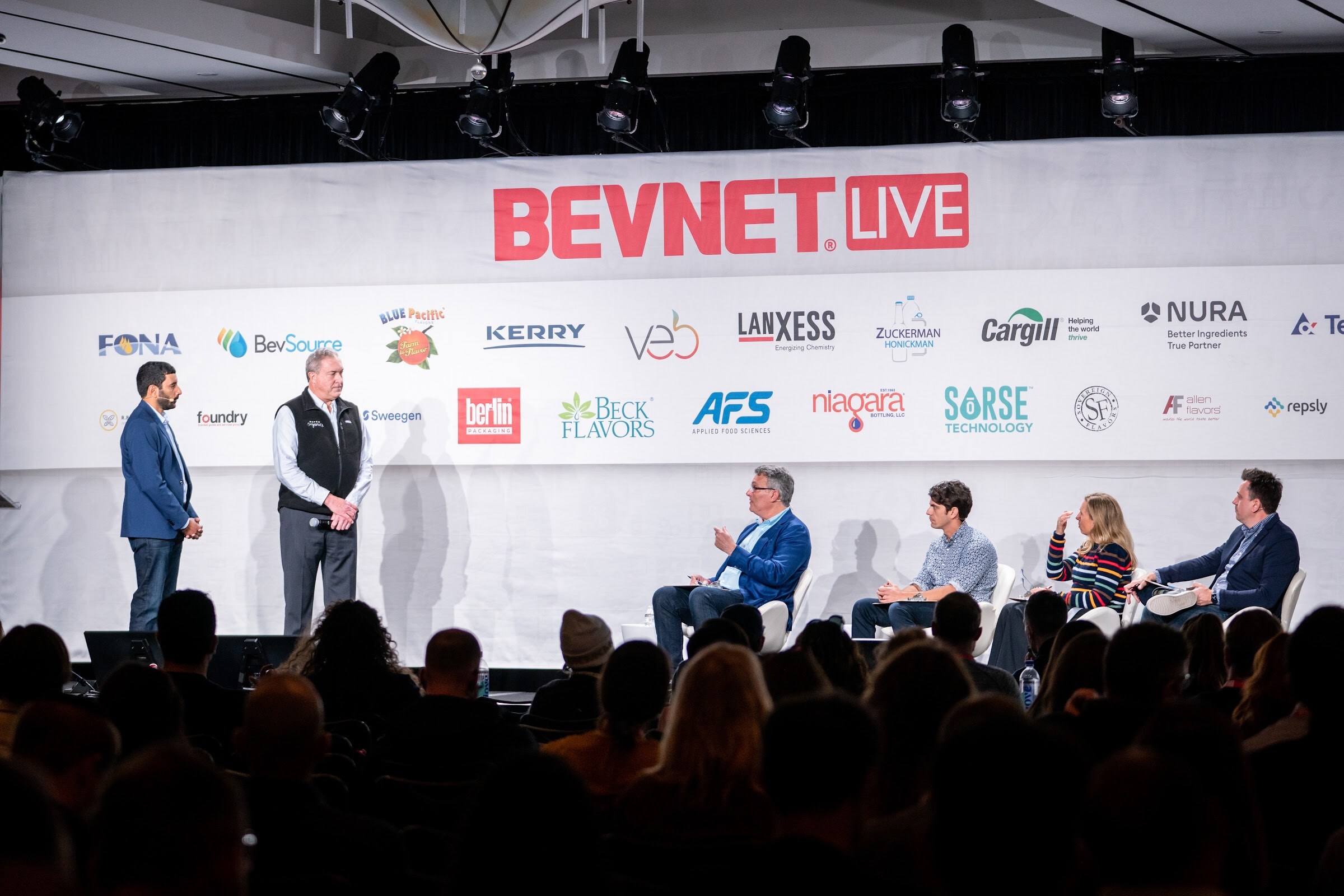 A wide view of a stage under a BevNET Live banner with two men standing at left and four judges seated at right in front of a silhouetted audience.