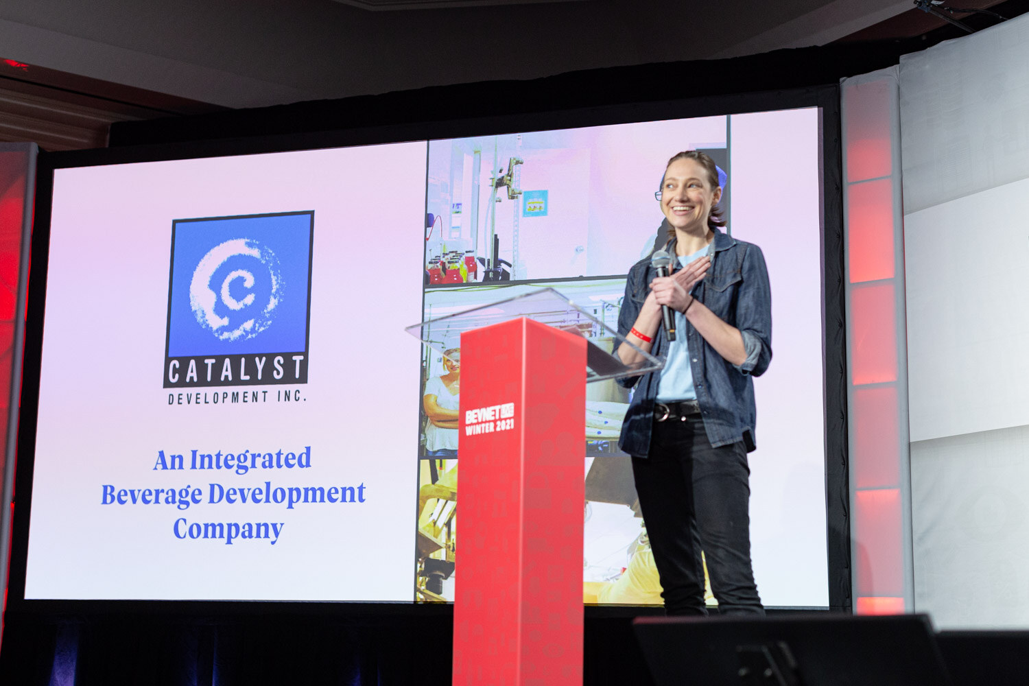 A woman in a denim jacket stands at a red podium beside a large projected slide with the Catalyst Development Inc. logo.