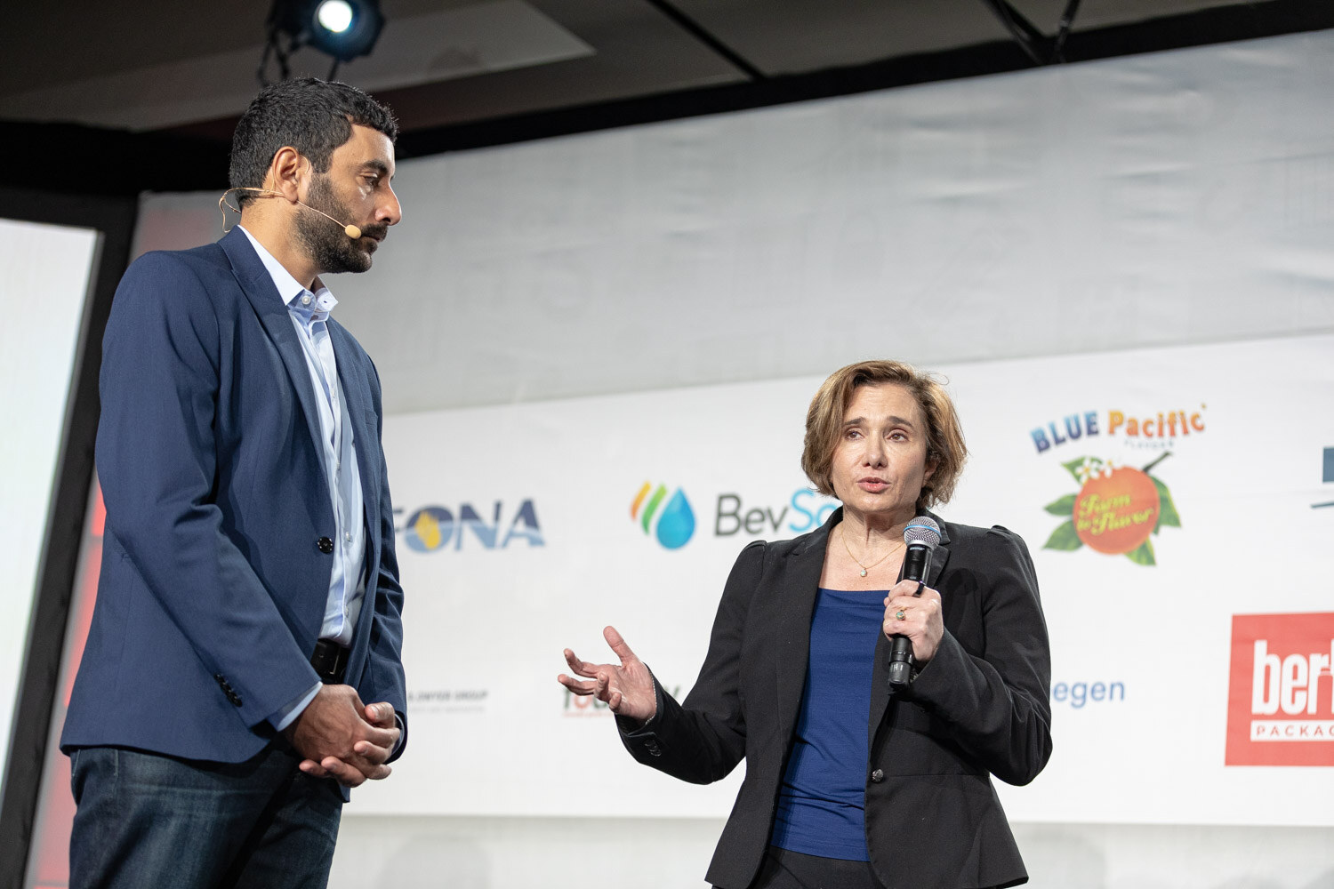 A man in a navy blazer stands with his hands clasped beside a woman in a black blazer who speaks into a microphone and gestures.