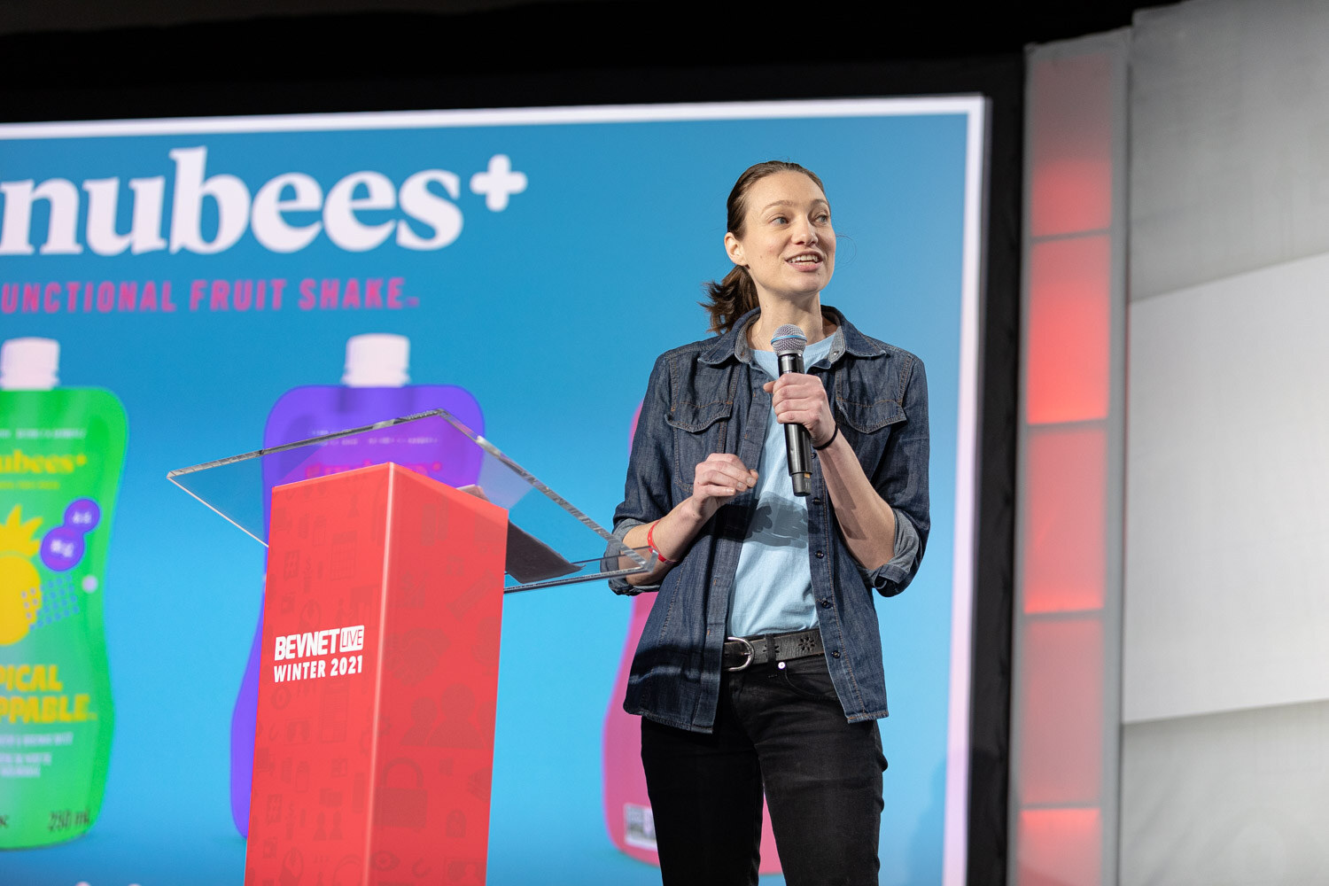 A woman in a denim jacket speaks into a microphone beside a red BevNET Live Winter 2021 podium in front of a large gnubees functional fruit shake slide.