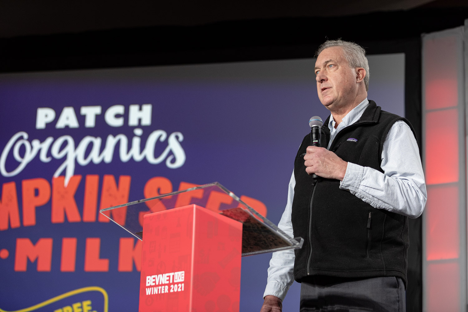 A gray-haired man in a black fleece vest holds a microphone at a red podium in front of a slide reading Patch Organics Pumpkin Seed Milk.