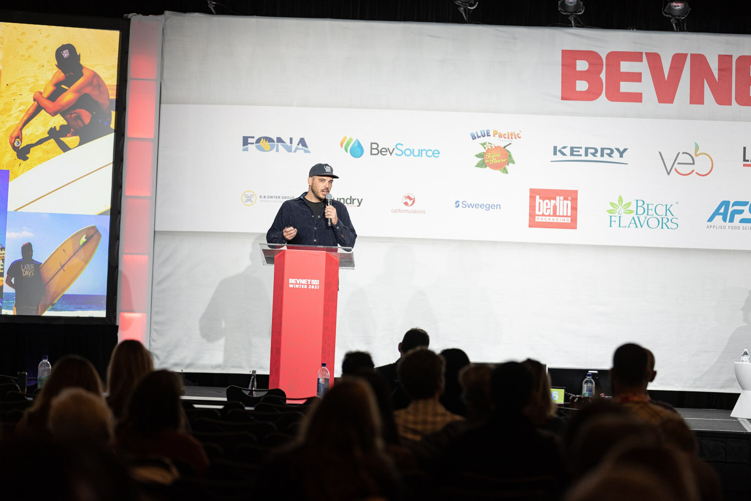 A man in a dark cap stands at a red podium on a stage with a surfer slide at left and a BevNET Live banner above, with the audience in front.