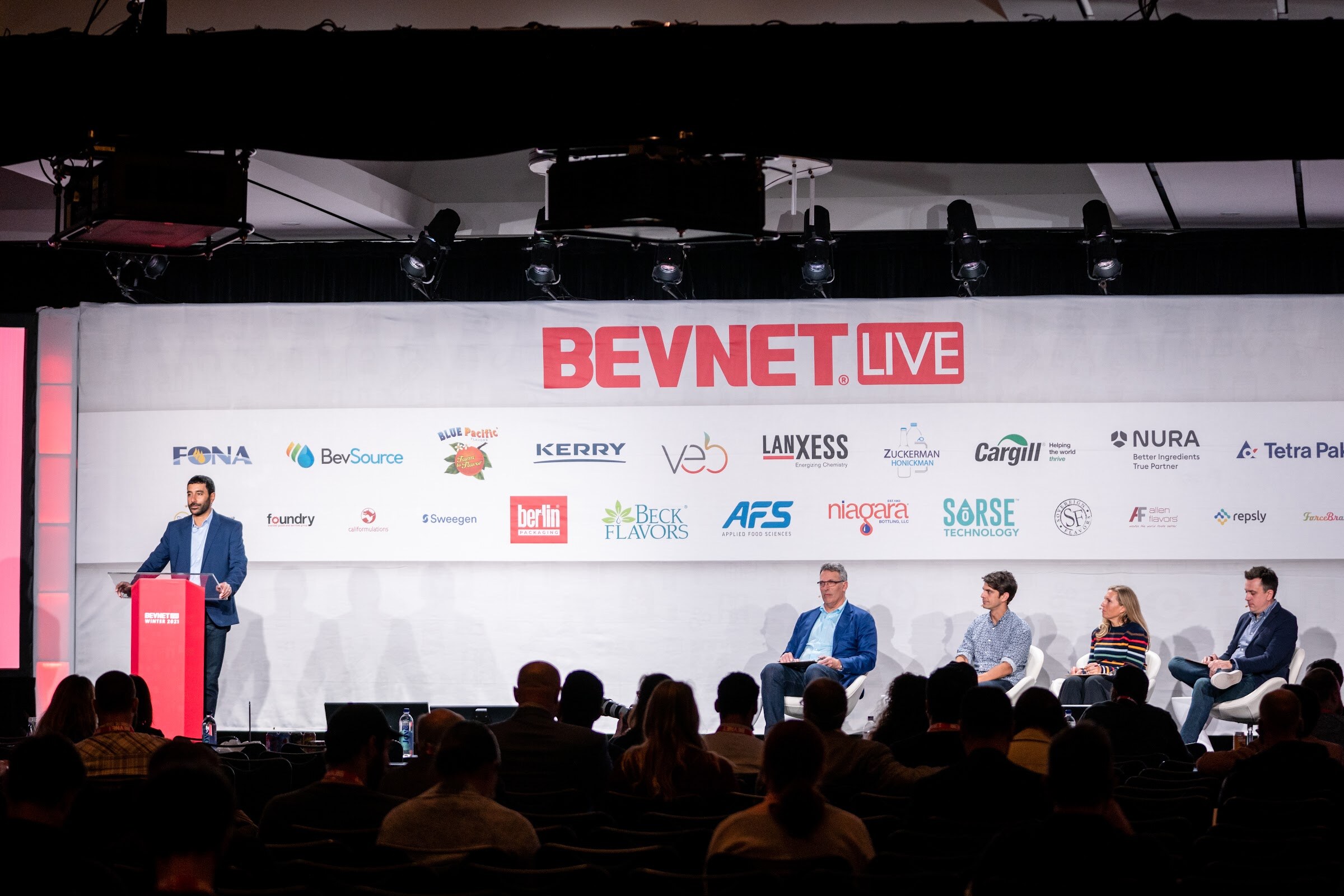 A wide view of a stage under a BevNET Live banner with a man at a red podium at left and four judges seated at right.