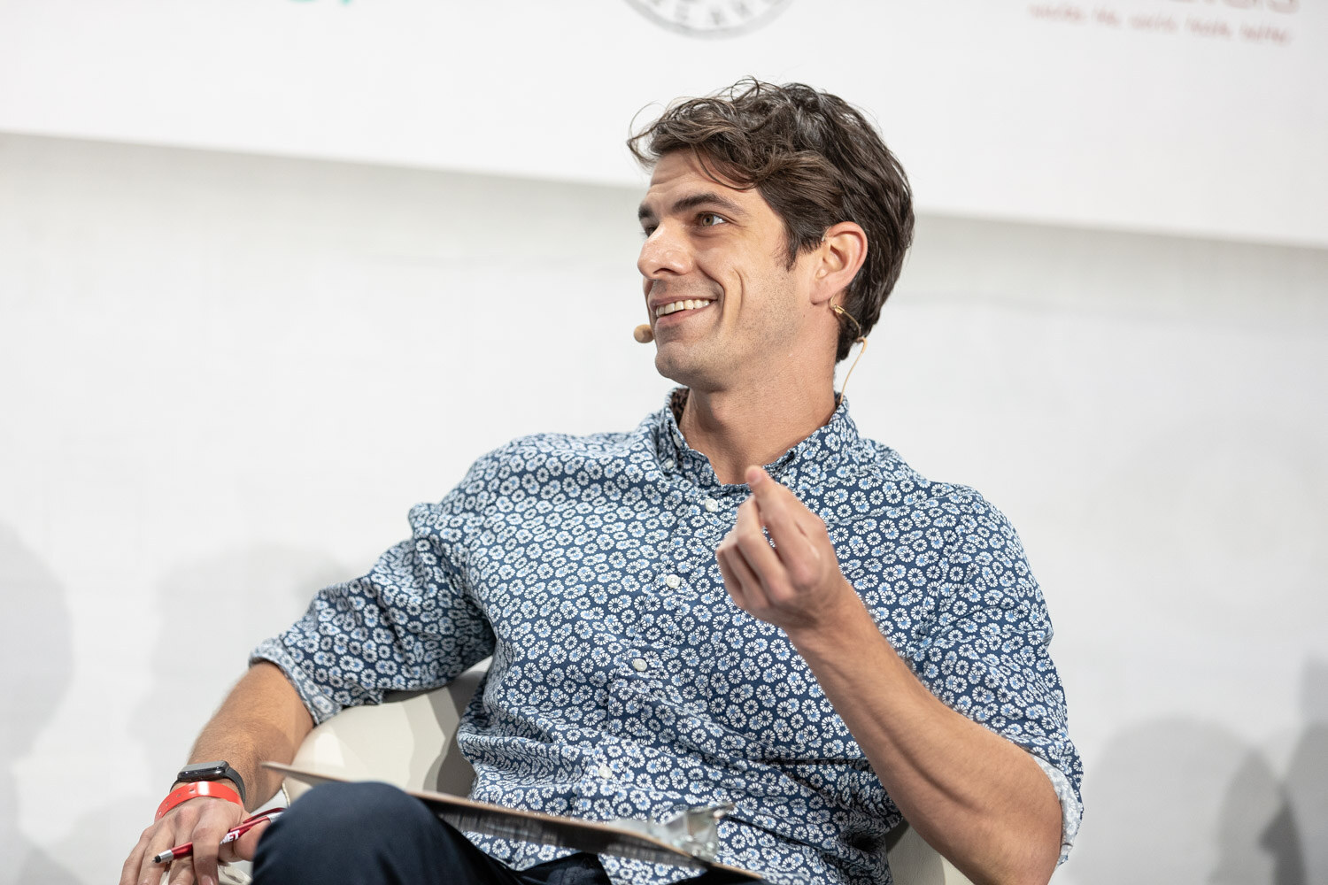 A dark-haired man in a blue patterned shirt with a clipboard and pen on his lap smiles and points a finger as he looks to the left.