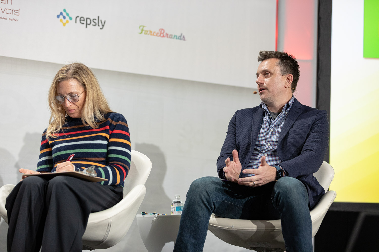 A blonde woman in a striped sweater writes on a clipboard next to a man in a navy blazer who gestures as he talks.