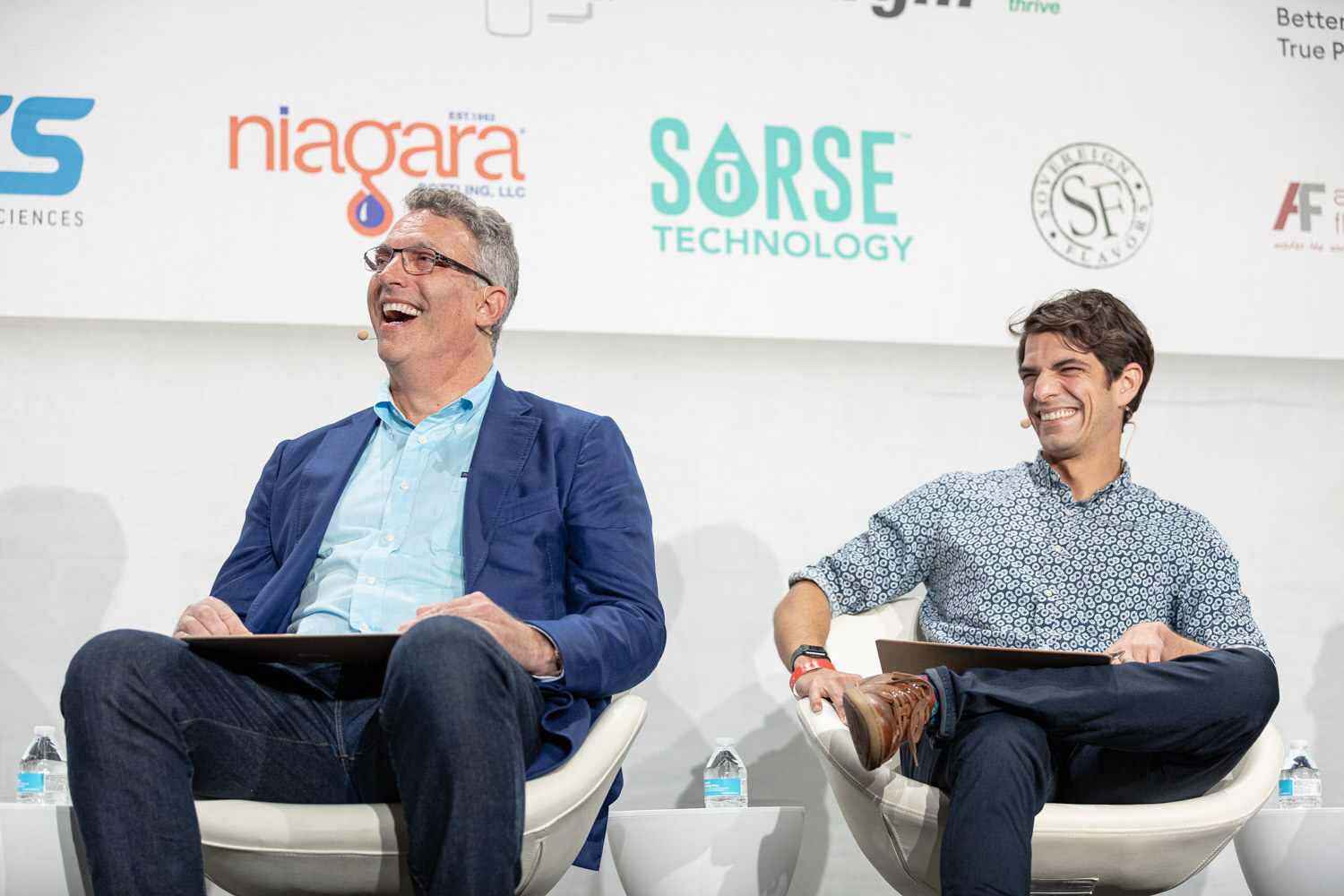 Two men in blue and patterned shirts seated in white chairs laugh, with sponsor logos including Niagara and Sorse Technology behind them.