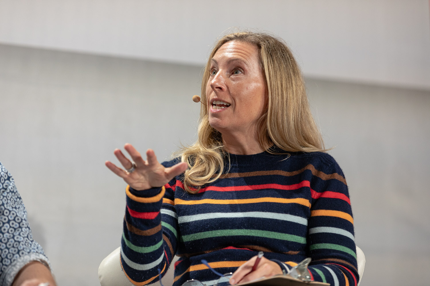 A blonde woman in a colorful striped sweater with a headset microphone raises her hand as she speaks.