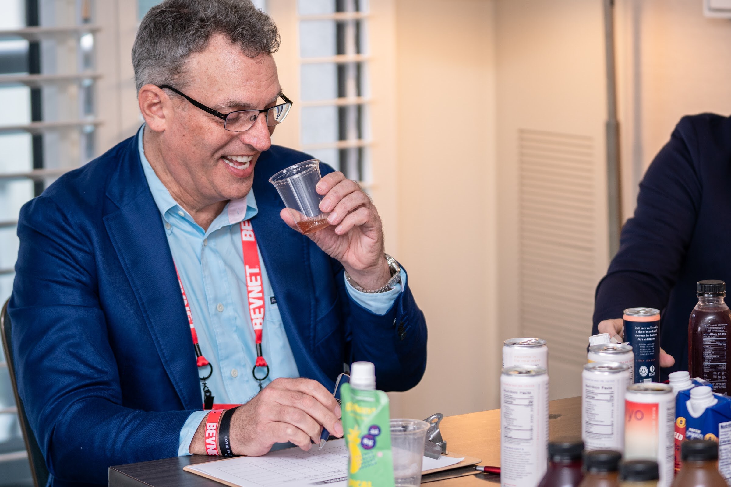 A man with glasses in a blue blazer and a BevNET lanyard sips from a small cup at a table with cans and a green pouch.