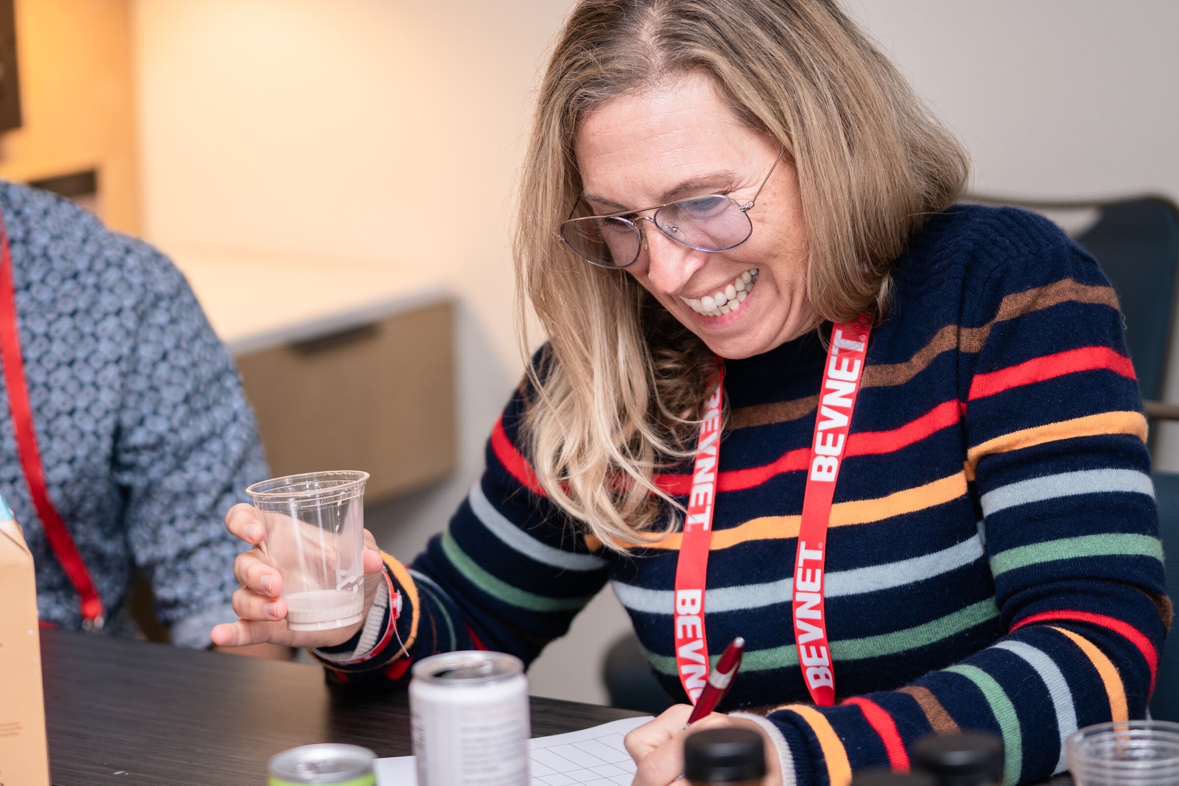 A woman with glasses and a BevNET lanyard holds a small cup and writes on a sheet at a table with cans.