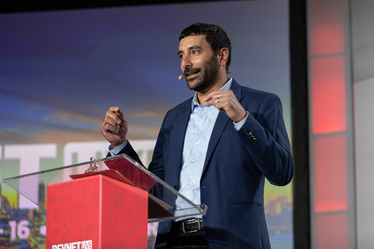 A man in a navy blazer and light blue shirt speaks and gestures at a red podium in front of a colorful screen.