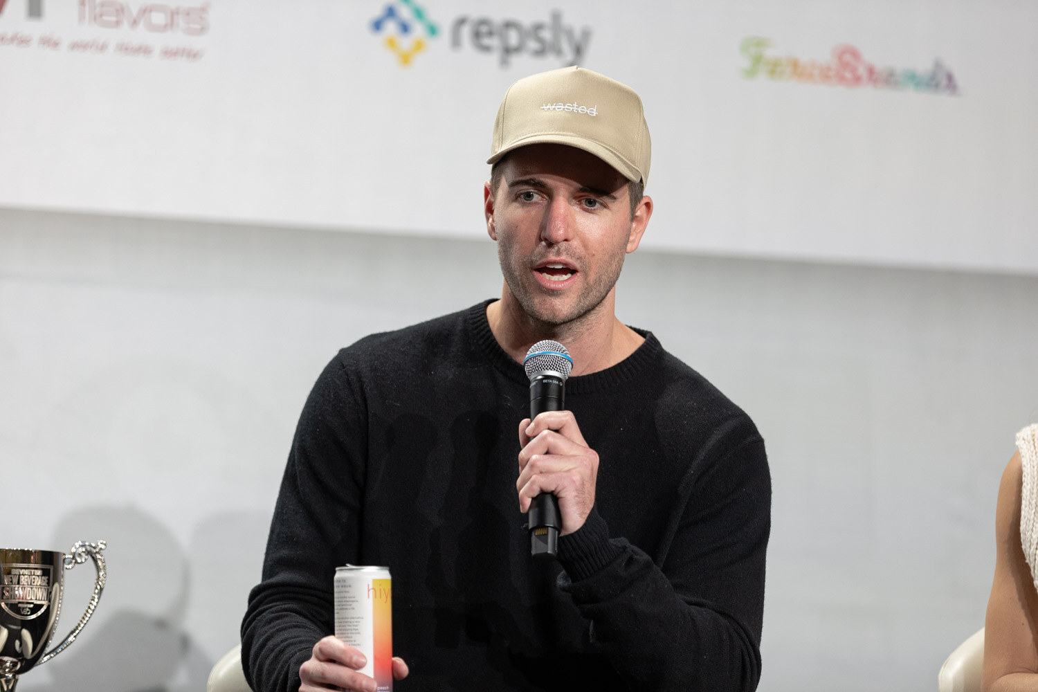 A man in a black sweater and tan cap holds a microphone and a can, with silver trophy cups visible at the edges.