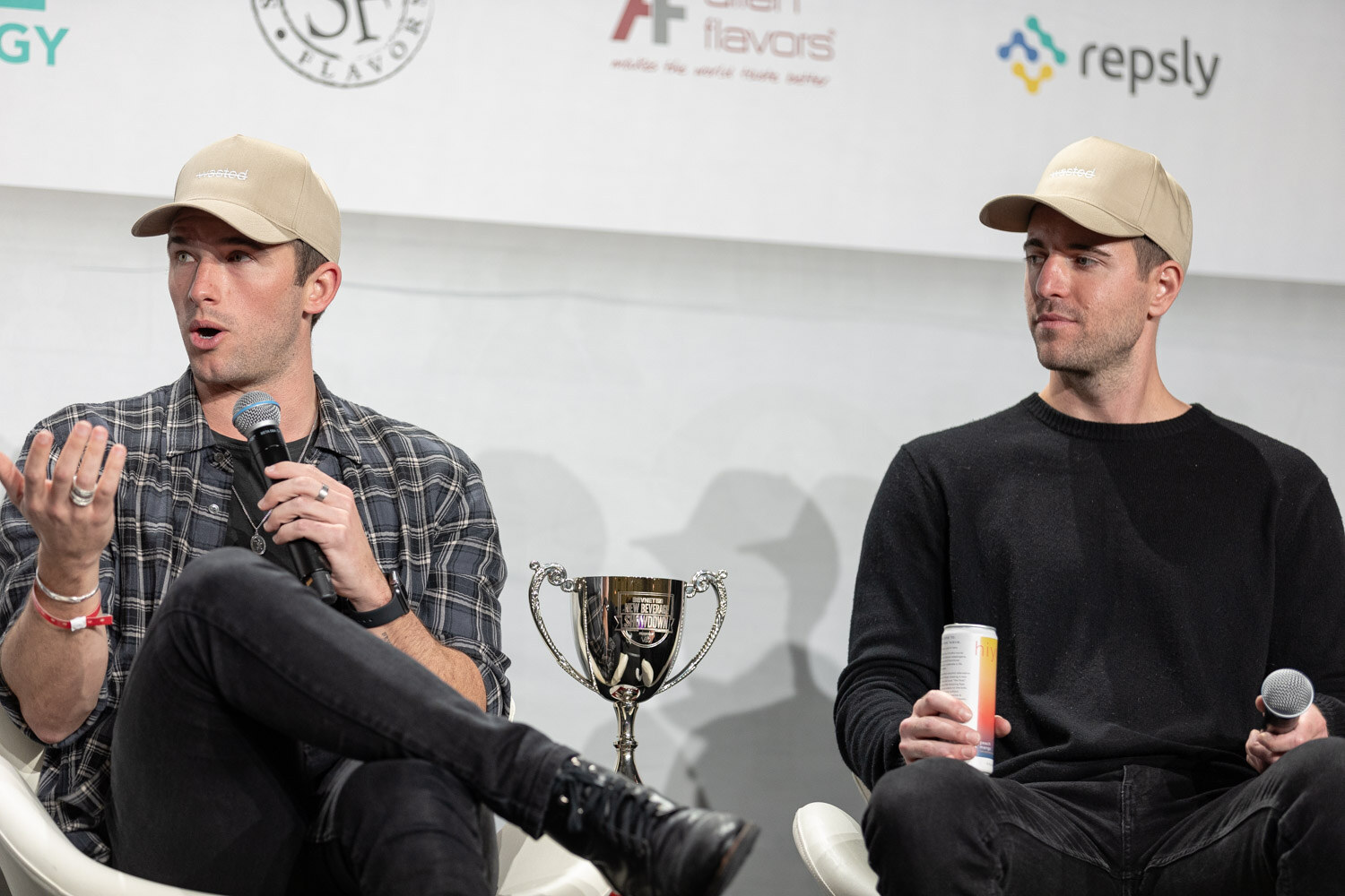A man in a plaid shirt gestures with a microphone while a man in a black sweater and tan cap looks on, with a silver trophy cup between them.