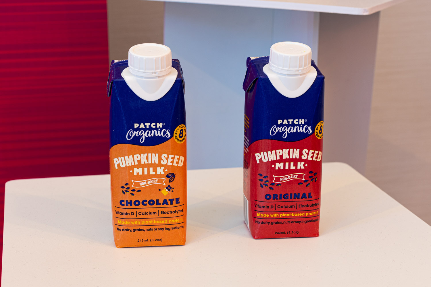 A close view of two cartons labeled Patch Organics Pumpkin Seed Milk, one orange Chocolate and one red Original, on a white table.